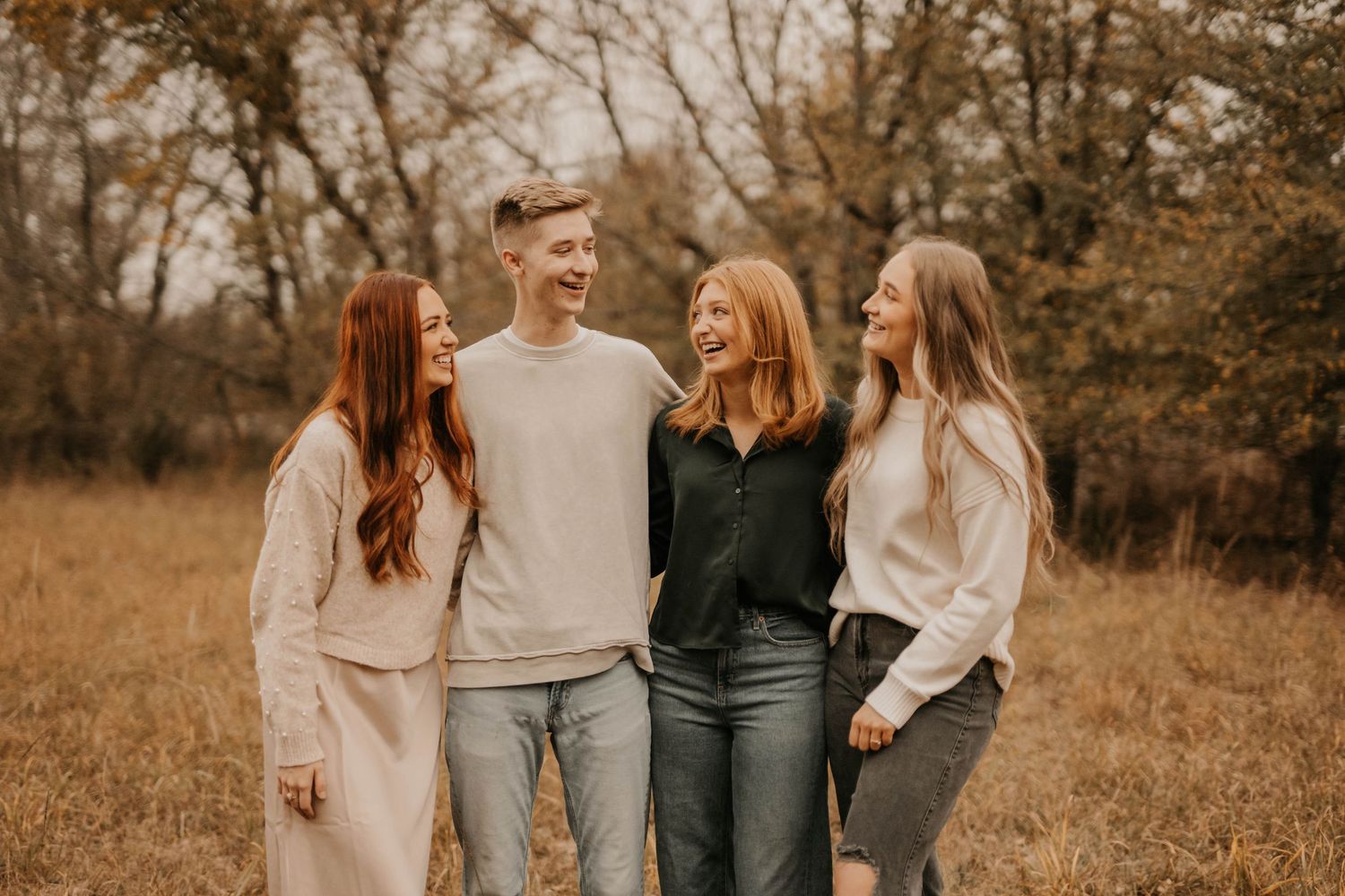 Group of friends in neutral colored outfits laughing together in fall field setting.