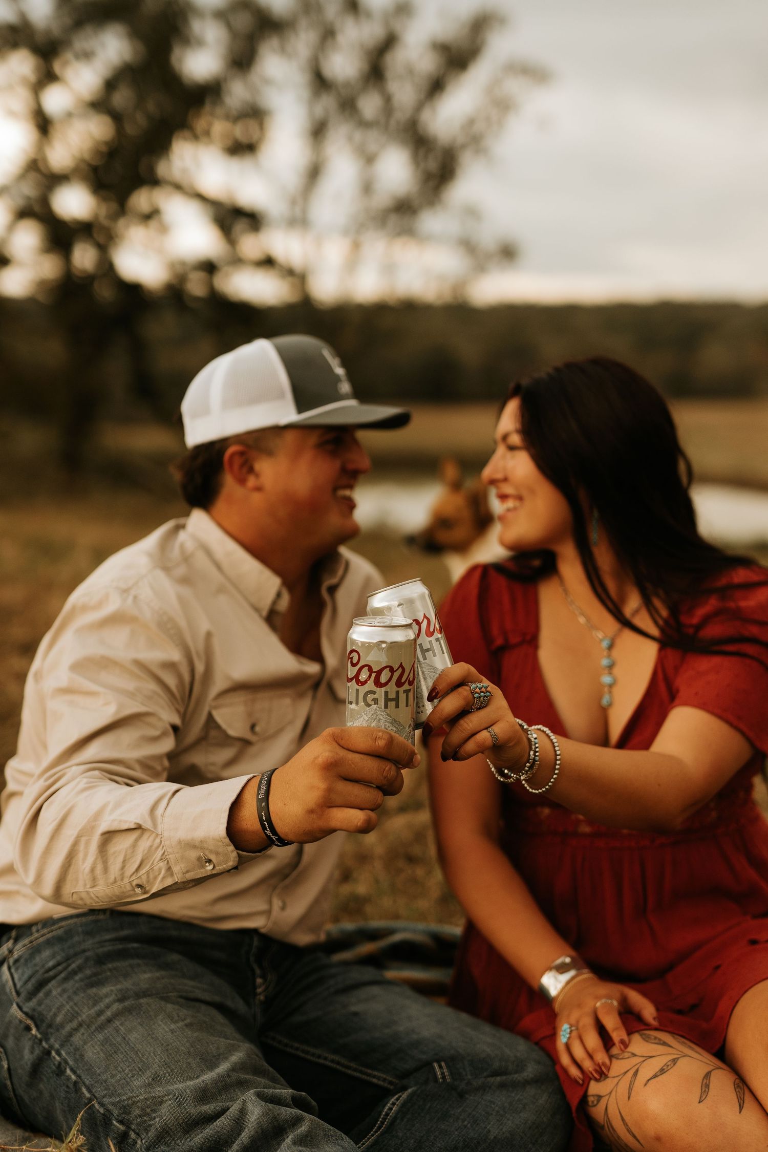 Couple toasting Coors Light cans outdoors at sunset, woman in red dress and man in white shirt and trucker hat.