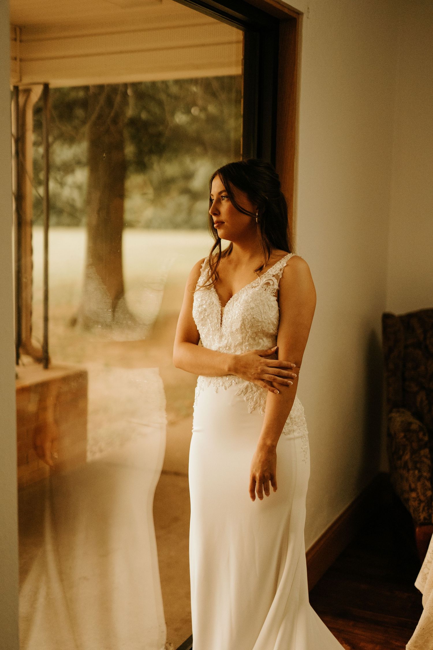 Bride in elegant white lace wedding gown standing by a glass door, gazing outside at lush greenery.