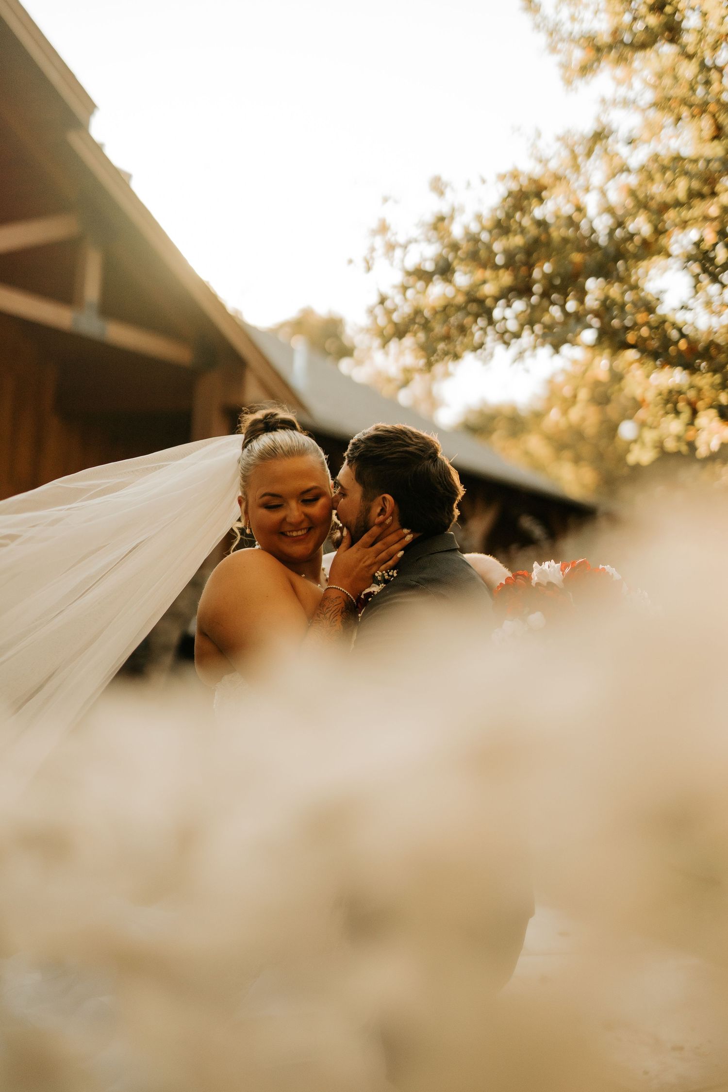 A joyful bride smiles as the groom leans in for a kiss outdoors, with a flowing veil and soft bokeh florals in the foreground.