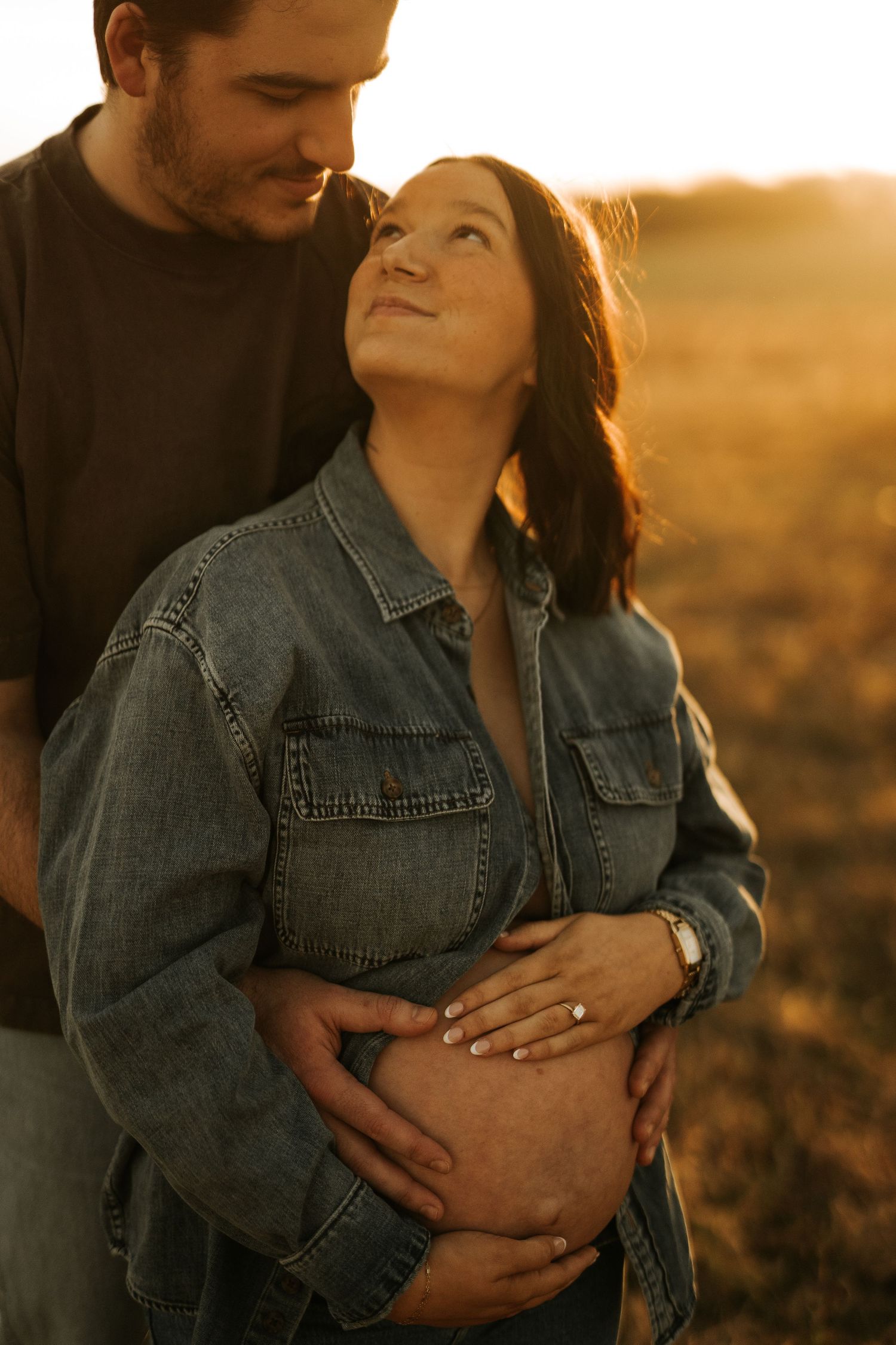 Couple embracing at golden hour sunset, pregnant woman in denim shirt cradling baby bump with partner in field.