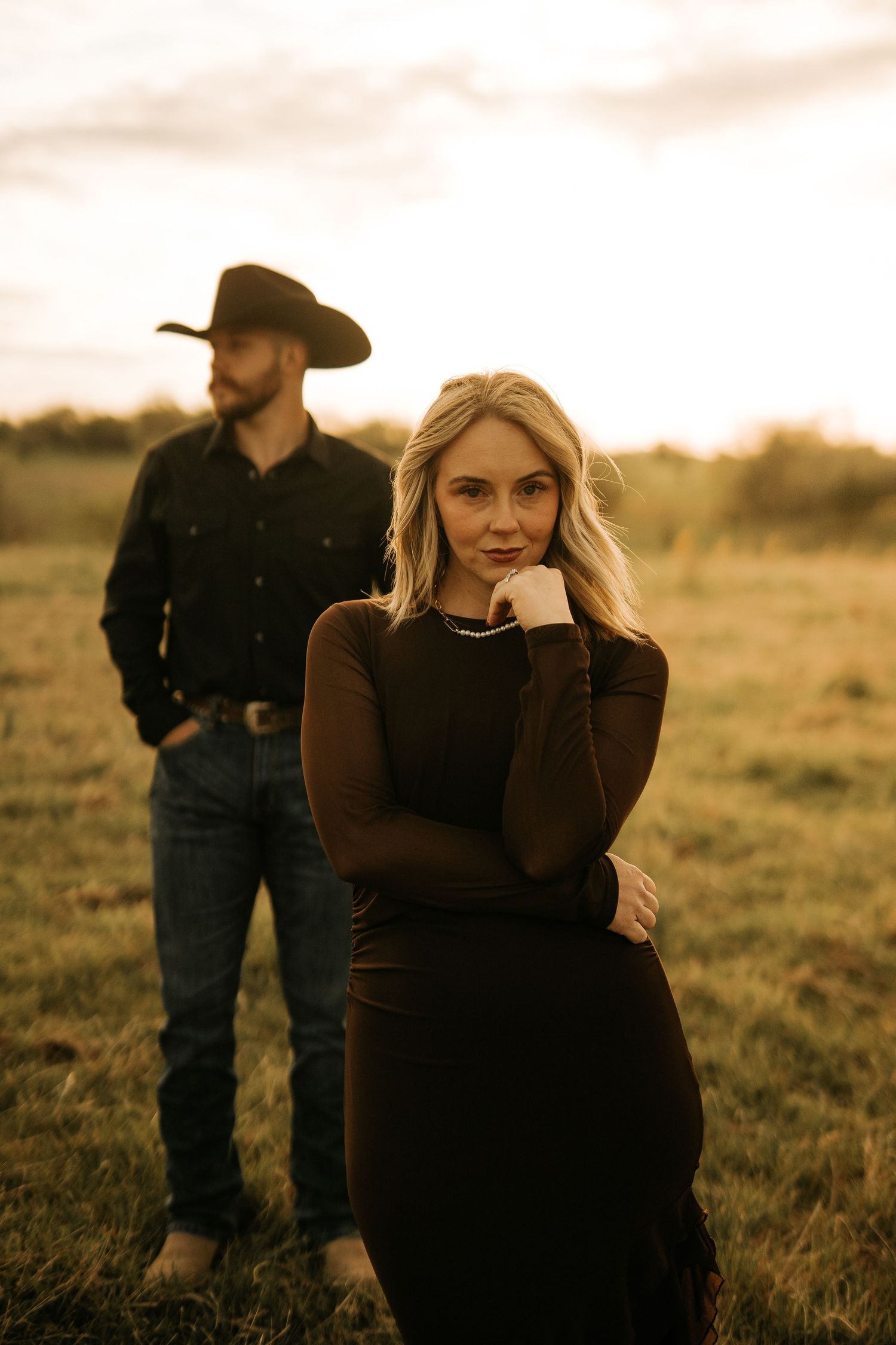 Blonde woman in black dress poses at golden hour with a man in cowboy hat standing in a field behind her.