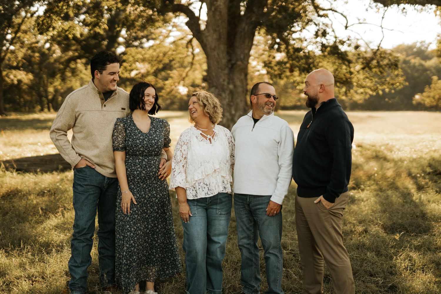 Five family members smiling and standing together outdoors near a large tree in a golden sunlit field.