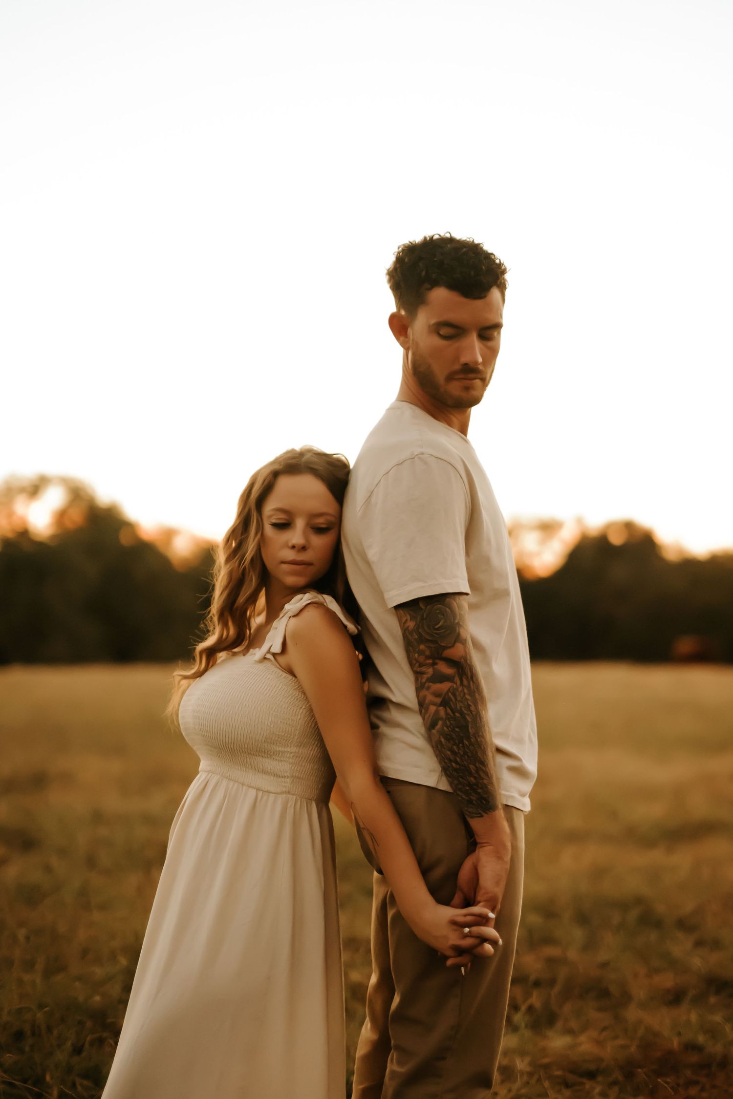 A couple stands back-to-back in a golden field at sunset, the man with a sleeve tattoo holding the woman's hand.