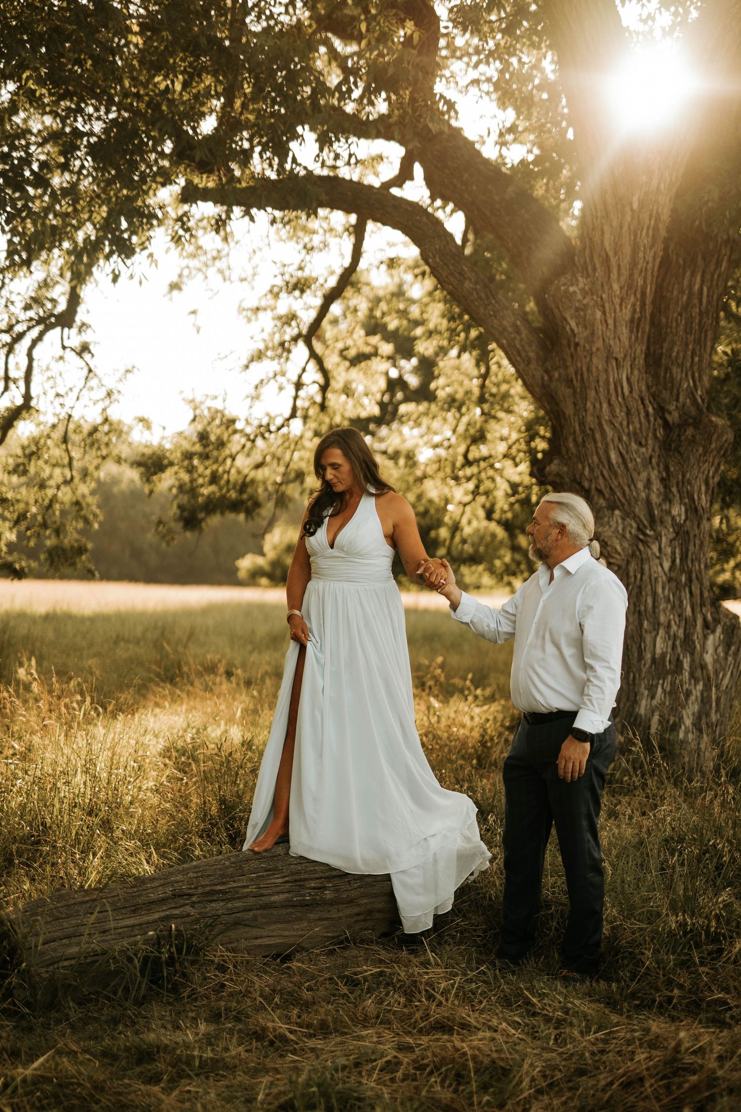 Bride in white halter gown stands on log at golden hour under oak tree as groom holds her hand in field.