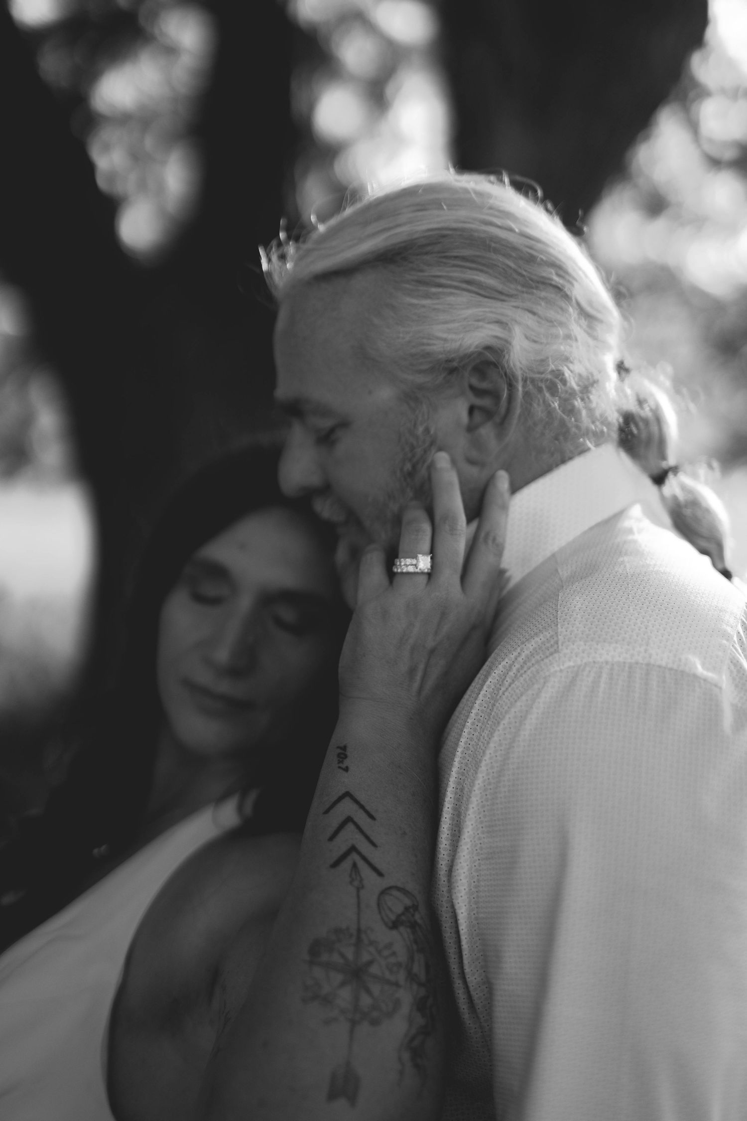 Black and white wedding photo of couple embracing, bride with tattoos tenderly touching grooms face outdoors.