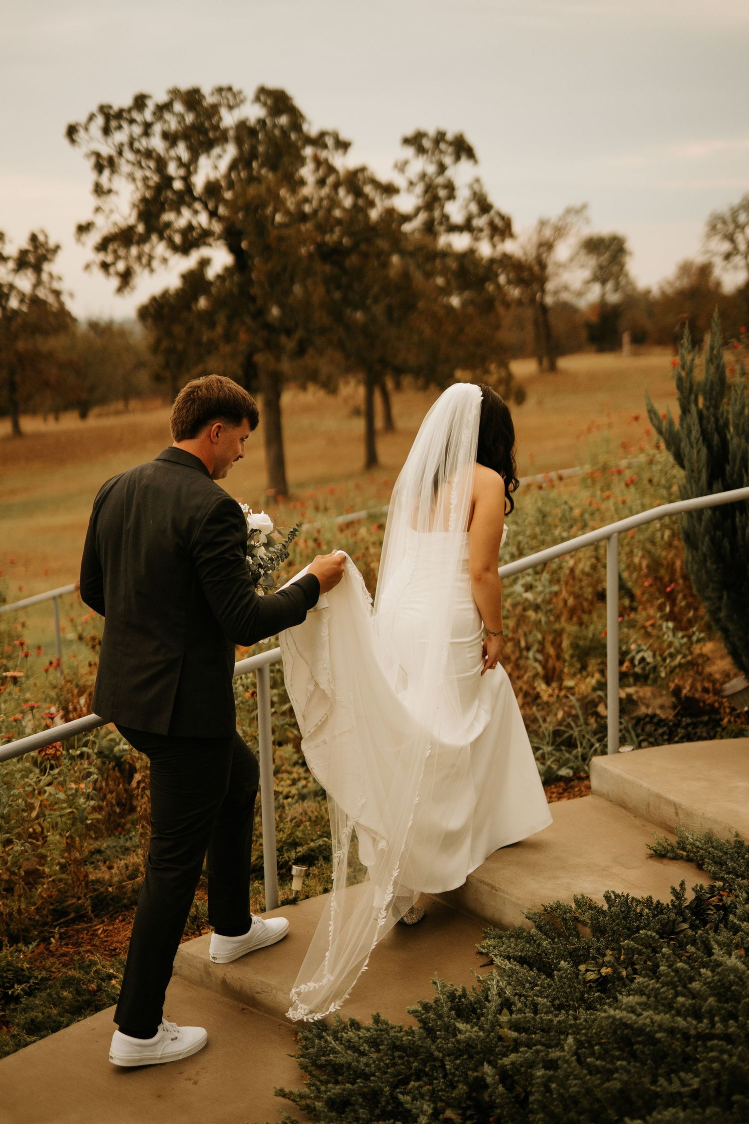 Groom in black suit holds bride's white gown train on outdoor steps at a fall wedding venue.