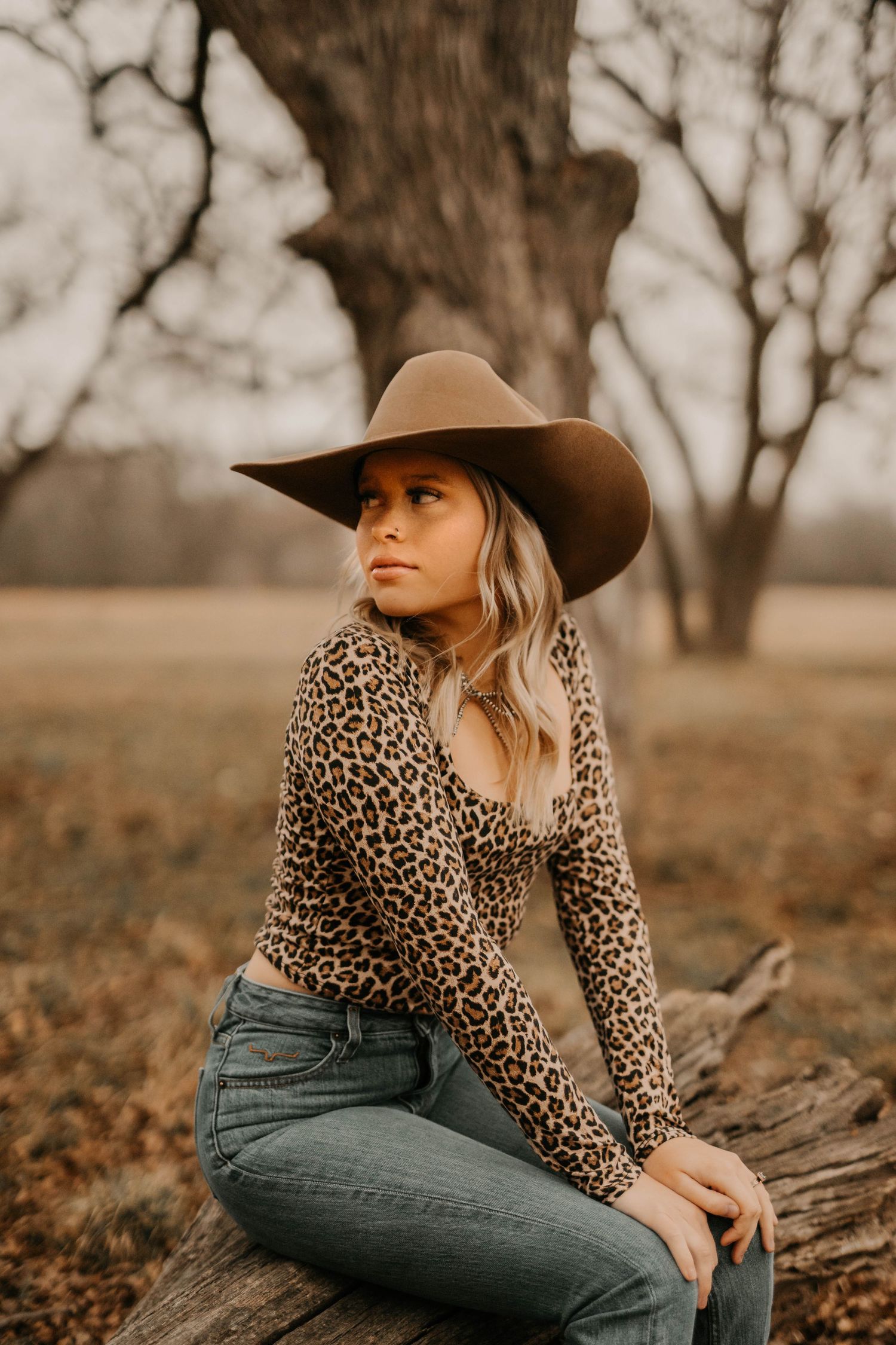 Woman in leopard print top and cowboy hat sits on log in rustic outdoor setting.