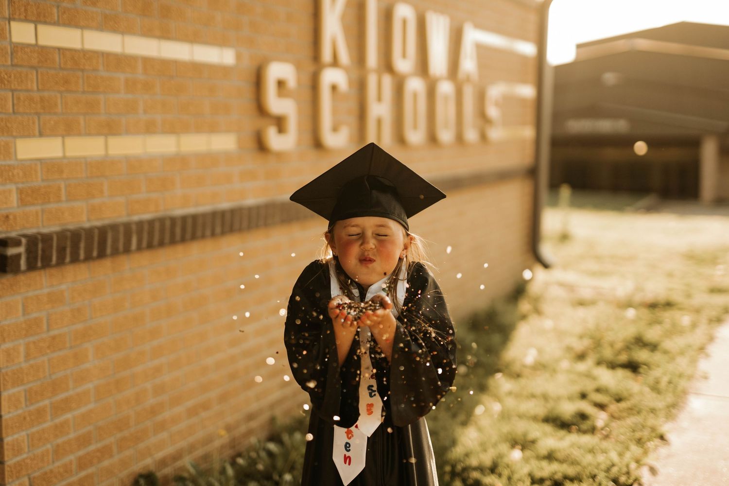 Graduate in cap and gown joyfully throws glitter outside Kiowa Schools.
