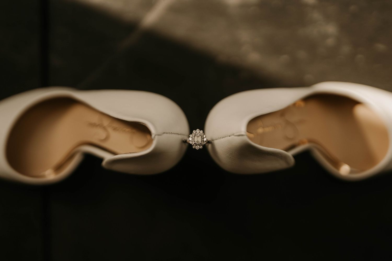 Artistic detail shot of white bridal shoes with diamond ring positioned between them.