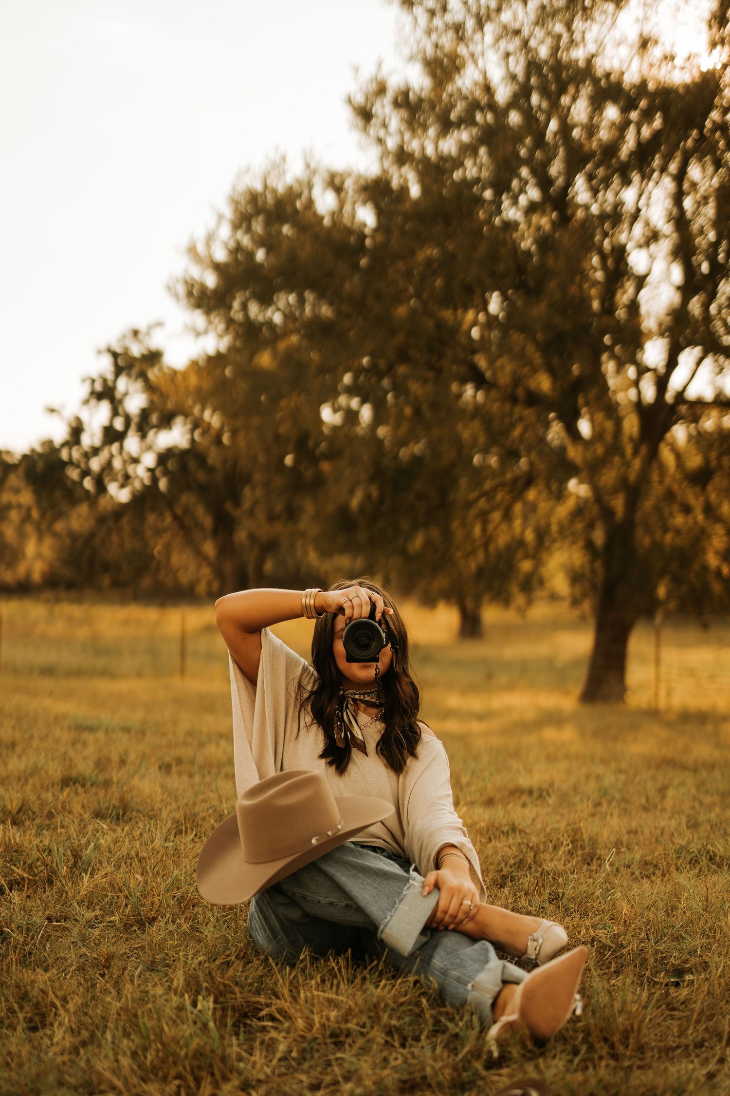 Woman sitting in golden field, holding camera up to face, wearing jeans and tan cardigan, trees in background.