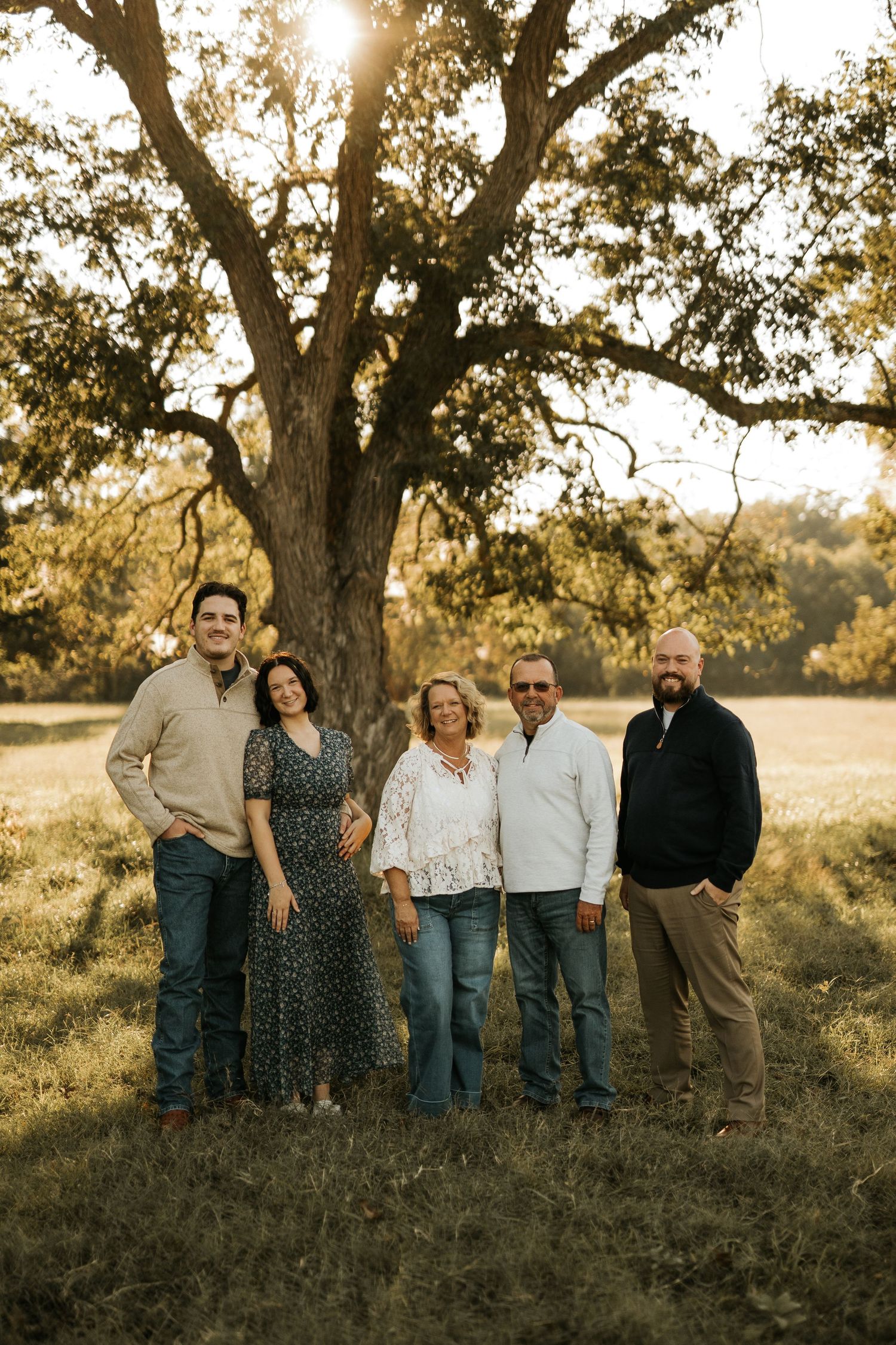 Family of five posing outdoors under a large oak tree during golden hour in a grassy field.