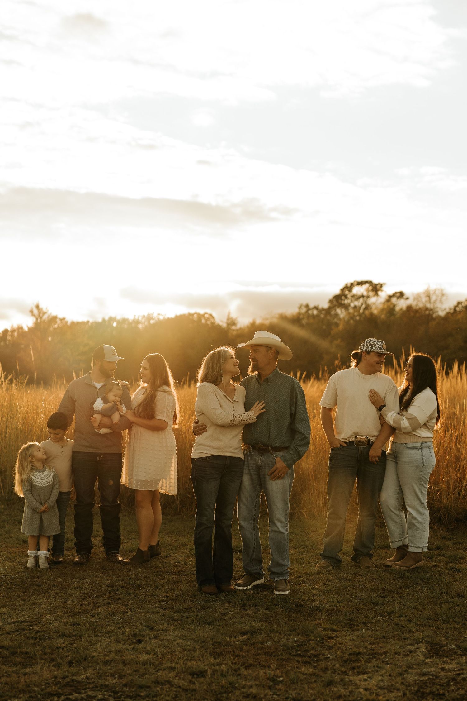 Family portrait at golden hour in a field, with couples embracing and children playing in warm sunset light.