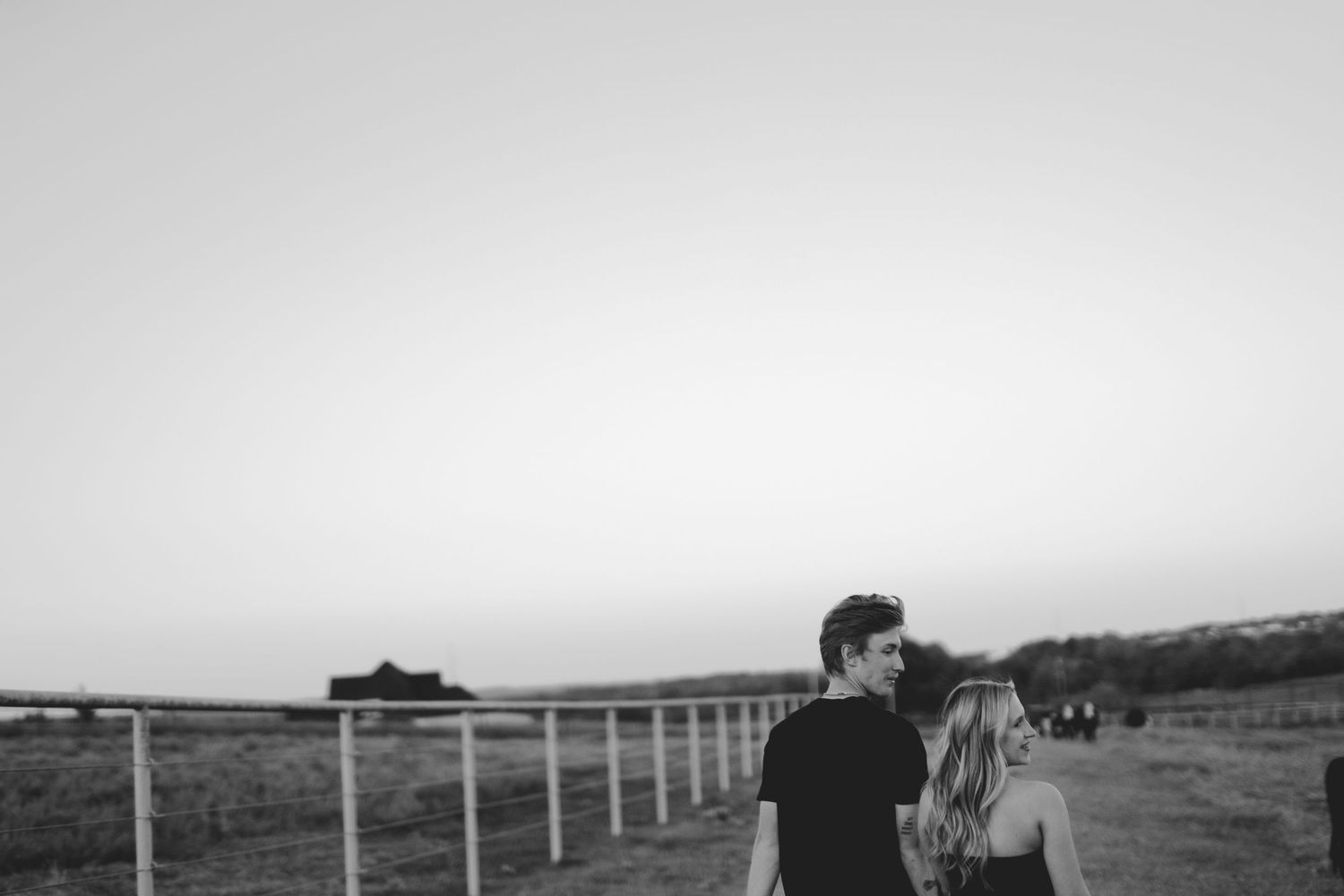 Black and white photo of a couple sharing a romantic moment outdoors near a fence at dusk.