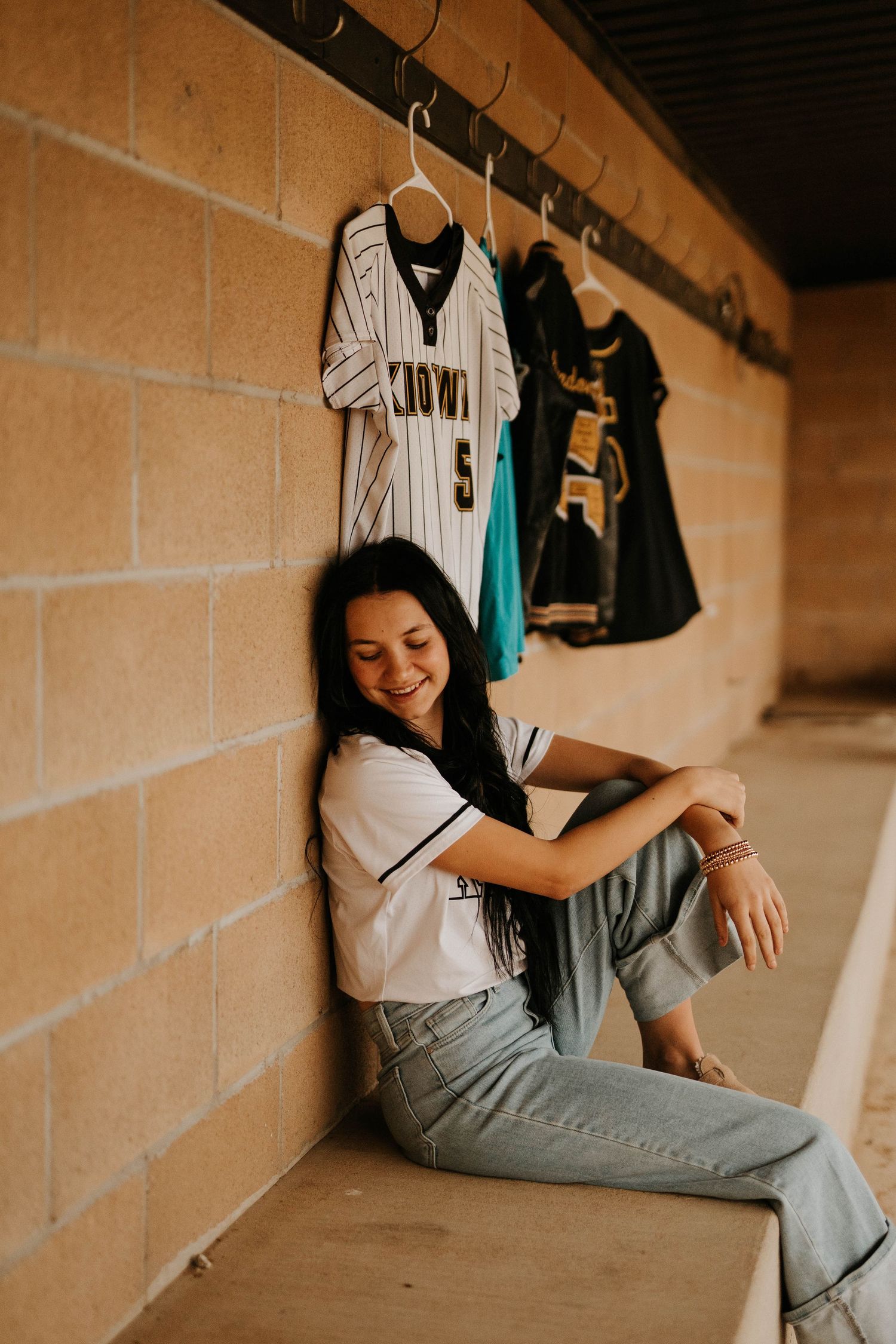 Person sits on floor of locker room with baseball jerseys hanging on wall.