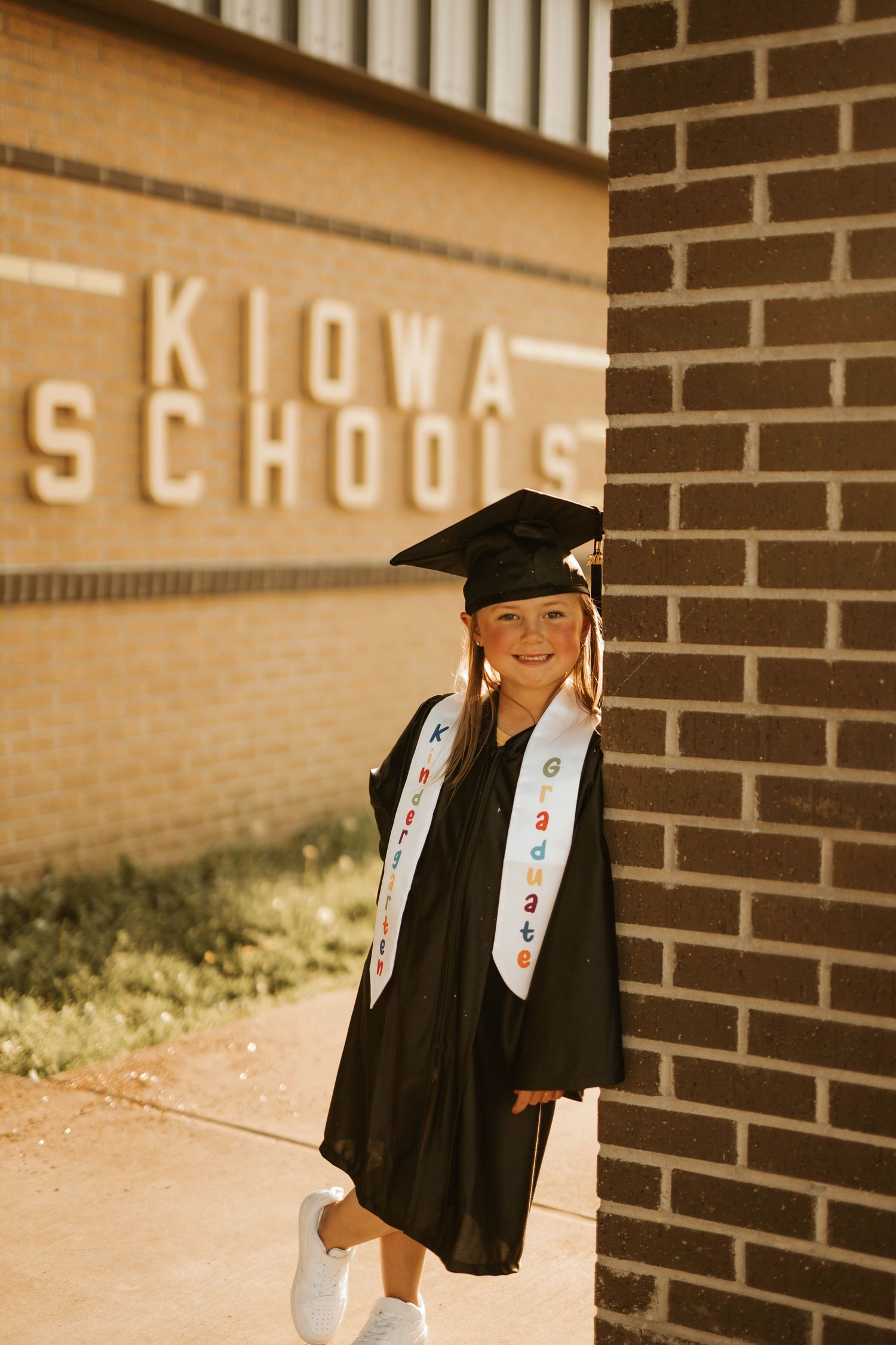 Young graduate in cap and gown poses outside Kiowa Schools building with decorative sash.