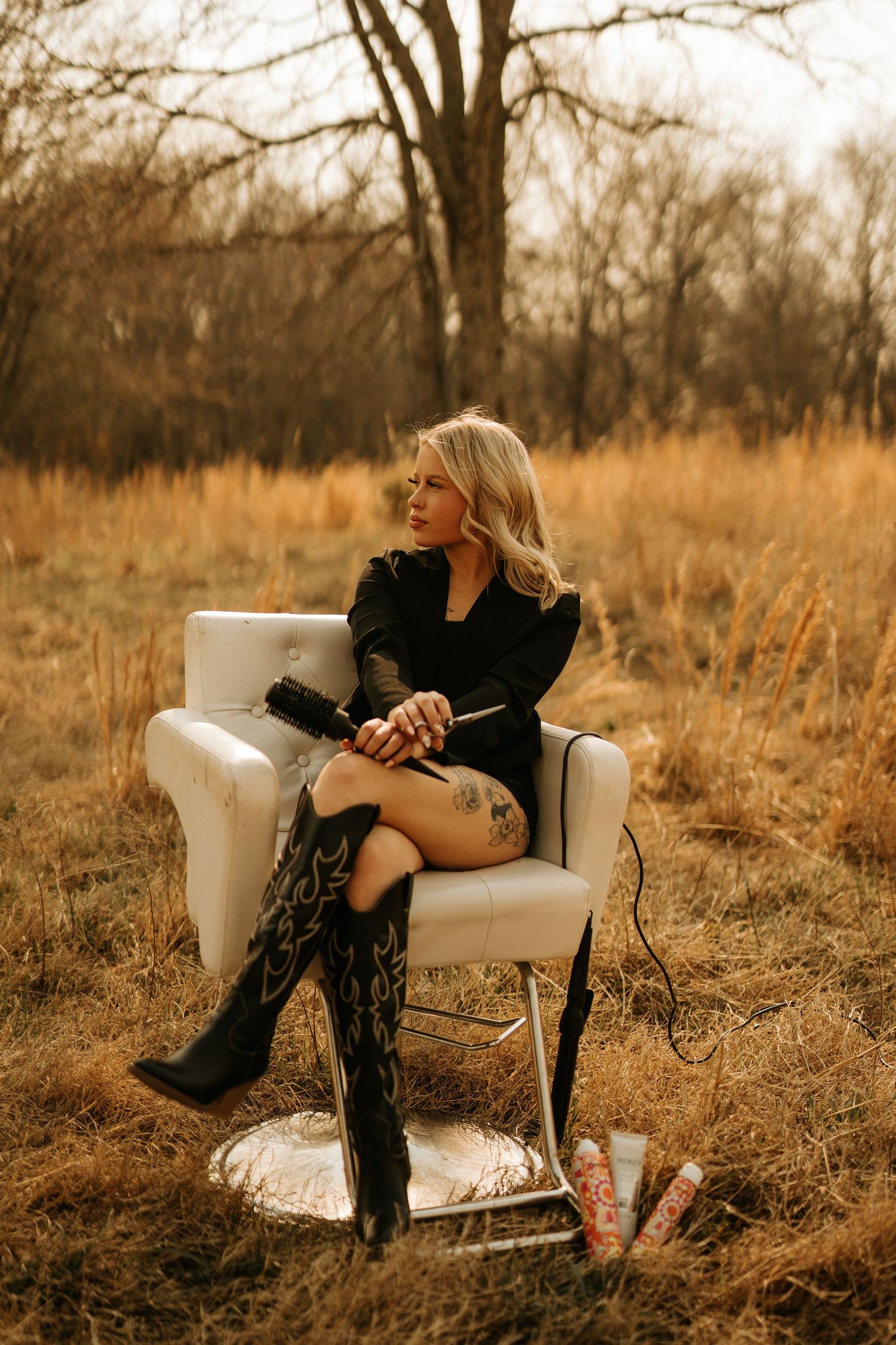 Blonde woman in black dress and cowboy boots sits in a salon chair outdoors in a golden field holding a hair dryer.