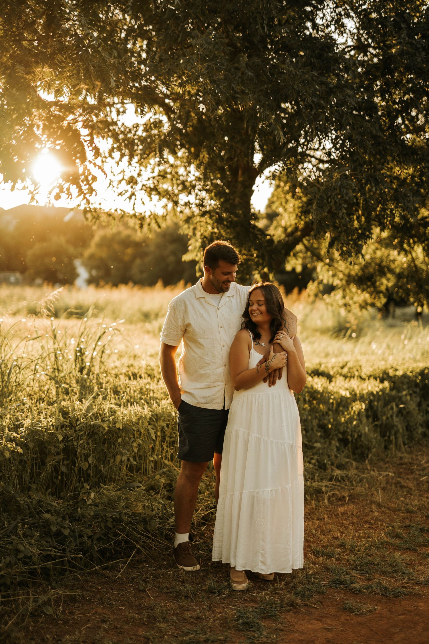 A couple embraces at golden hour in a sunlit field, the woman in a white maxi dress, standing near a large tree.