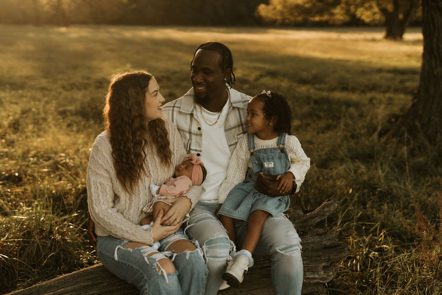 A multiracial family sits outdoors at golden hour, parents smiling at each other while holding a newborn and toddler daughter.