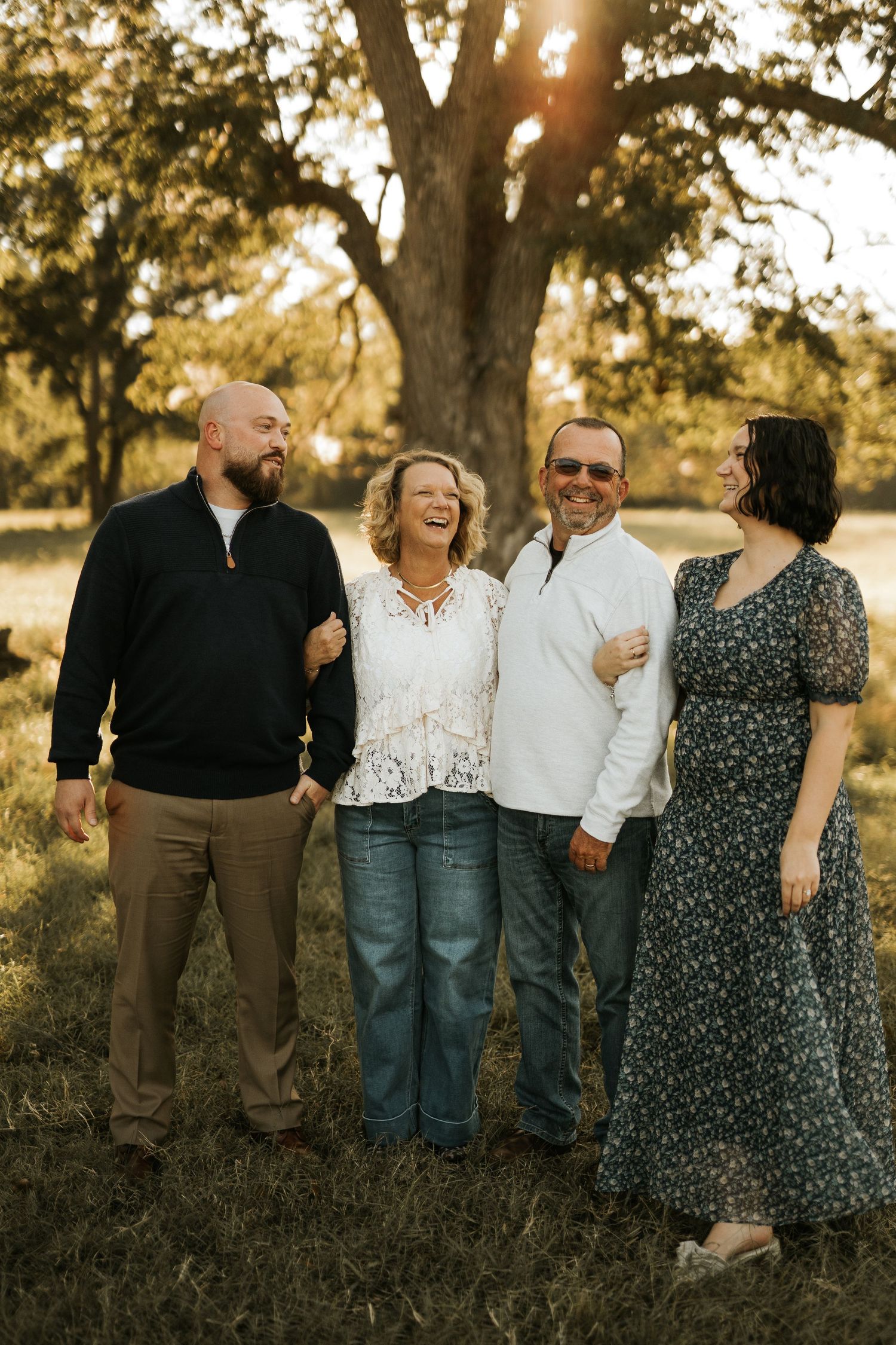 Four smiling family members pose outdoors at golden hour beneath a large oak tree in casual attire.
