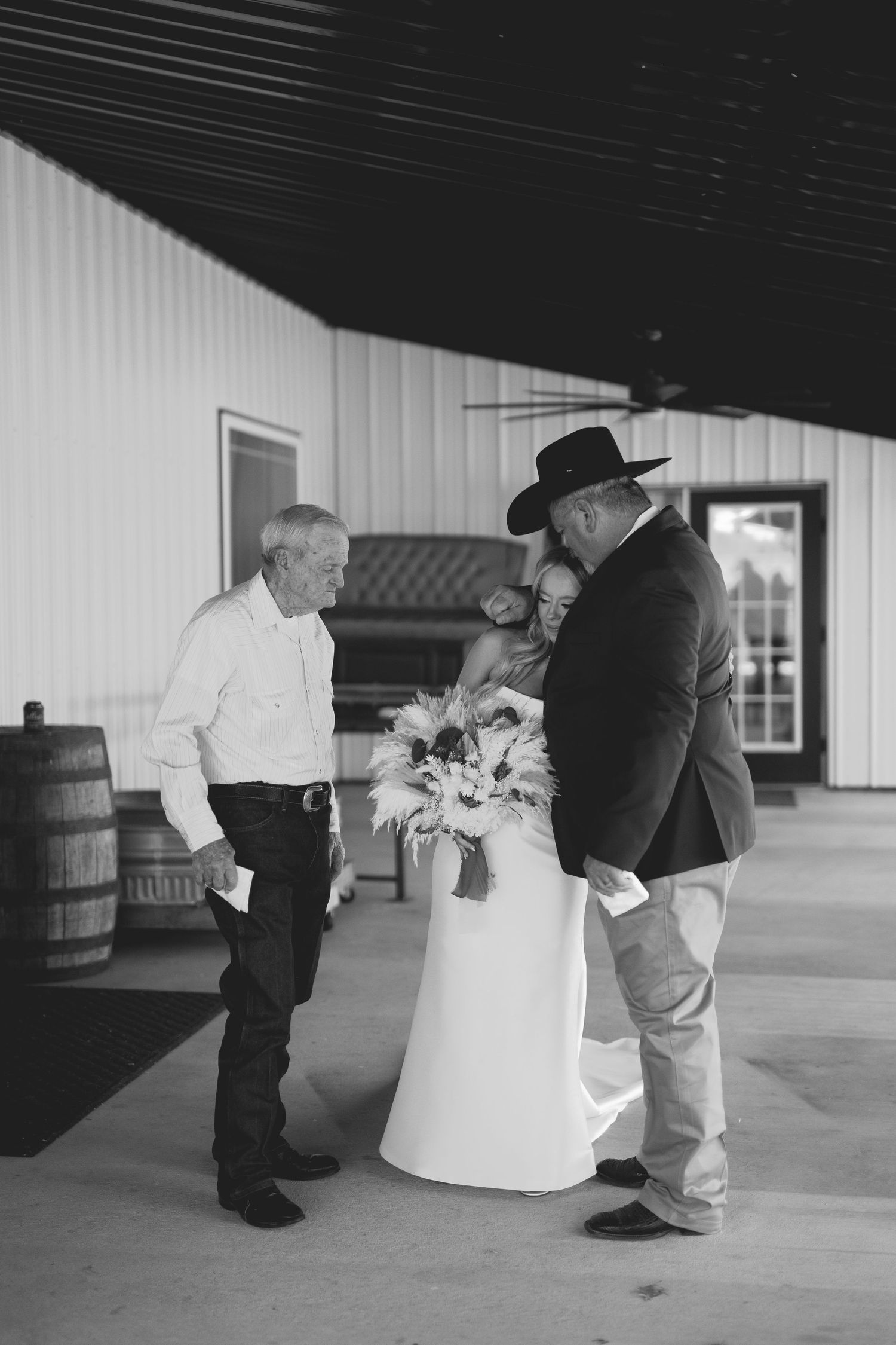 Black and white wedding photo of bride and groom embracing while holding bouquet, with elderly guest watching at rustic barn venue.