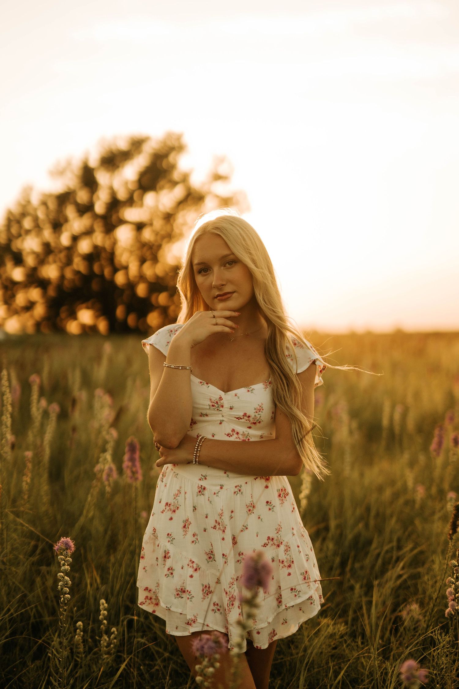 Blonde woman in floral white dress standing in a sunlit field of wildflowers during golden hour sunset.