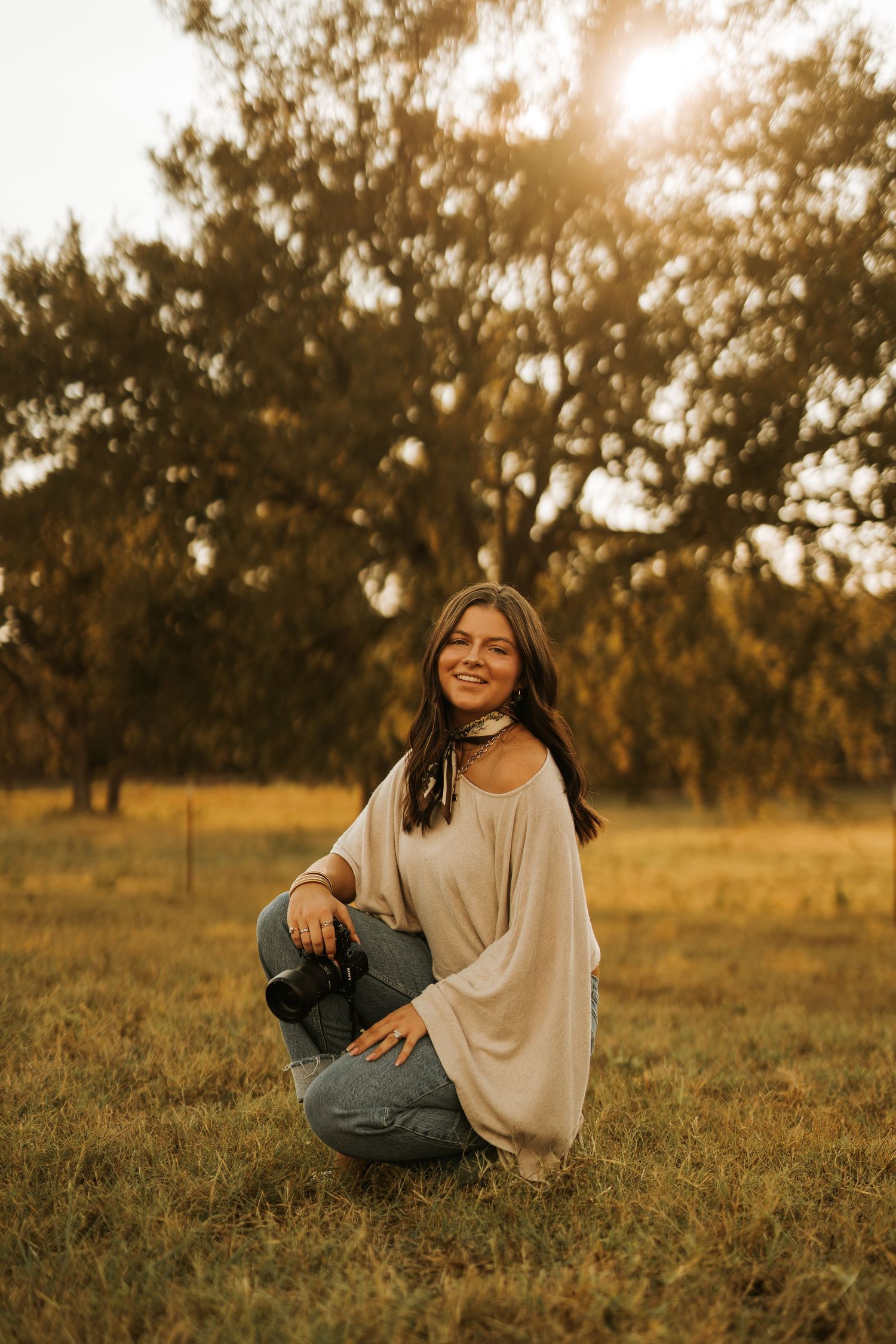 Young woman smiling in golden hour sunlight, holding camera, sitting in grassy field with trees in background.