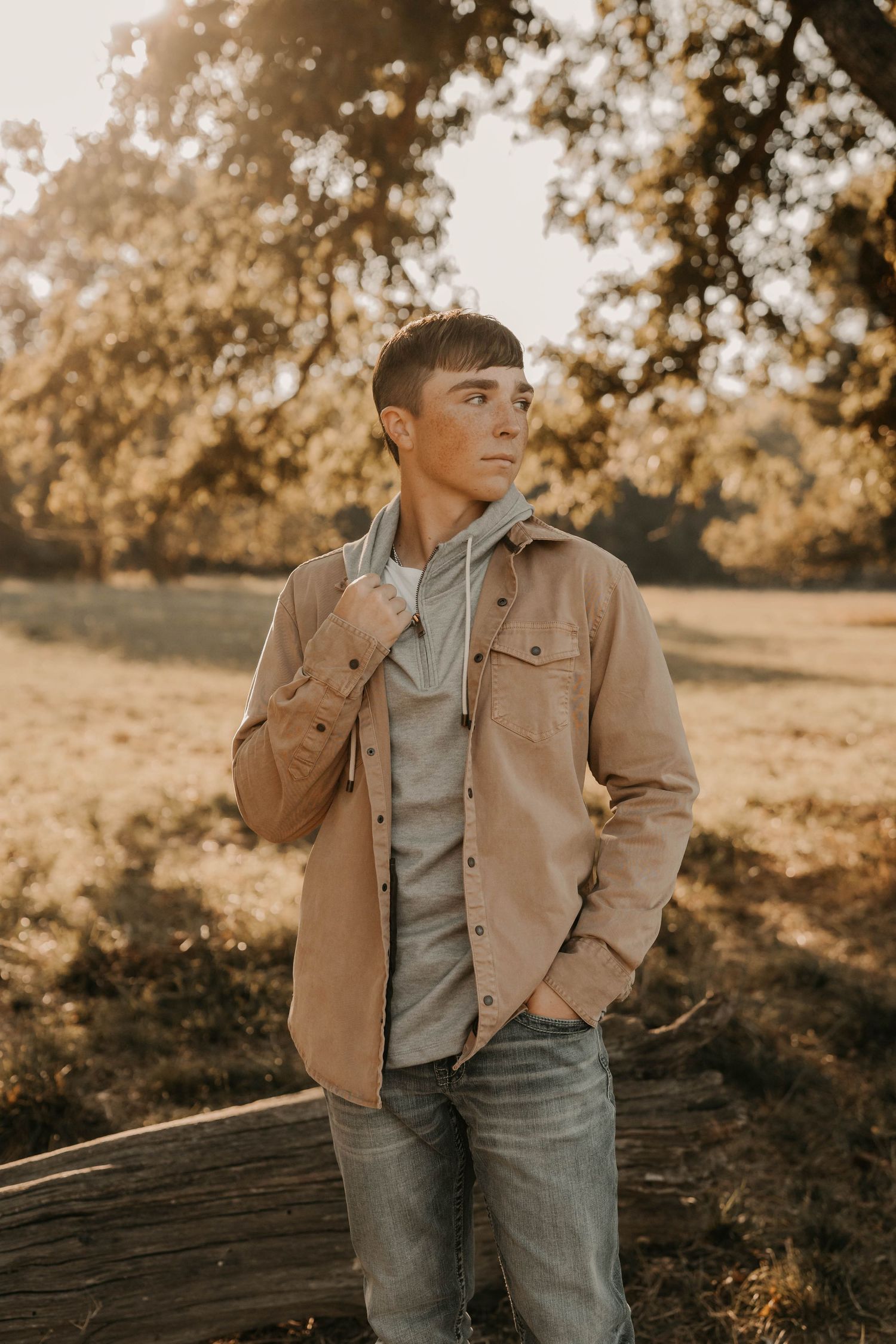 Person wearing tan jacket and grey hoodie poses against autumn trees at sunset.