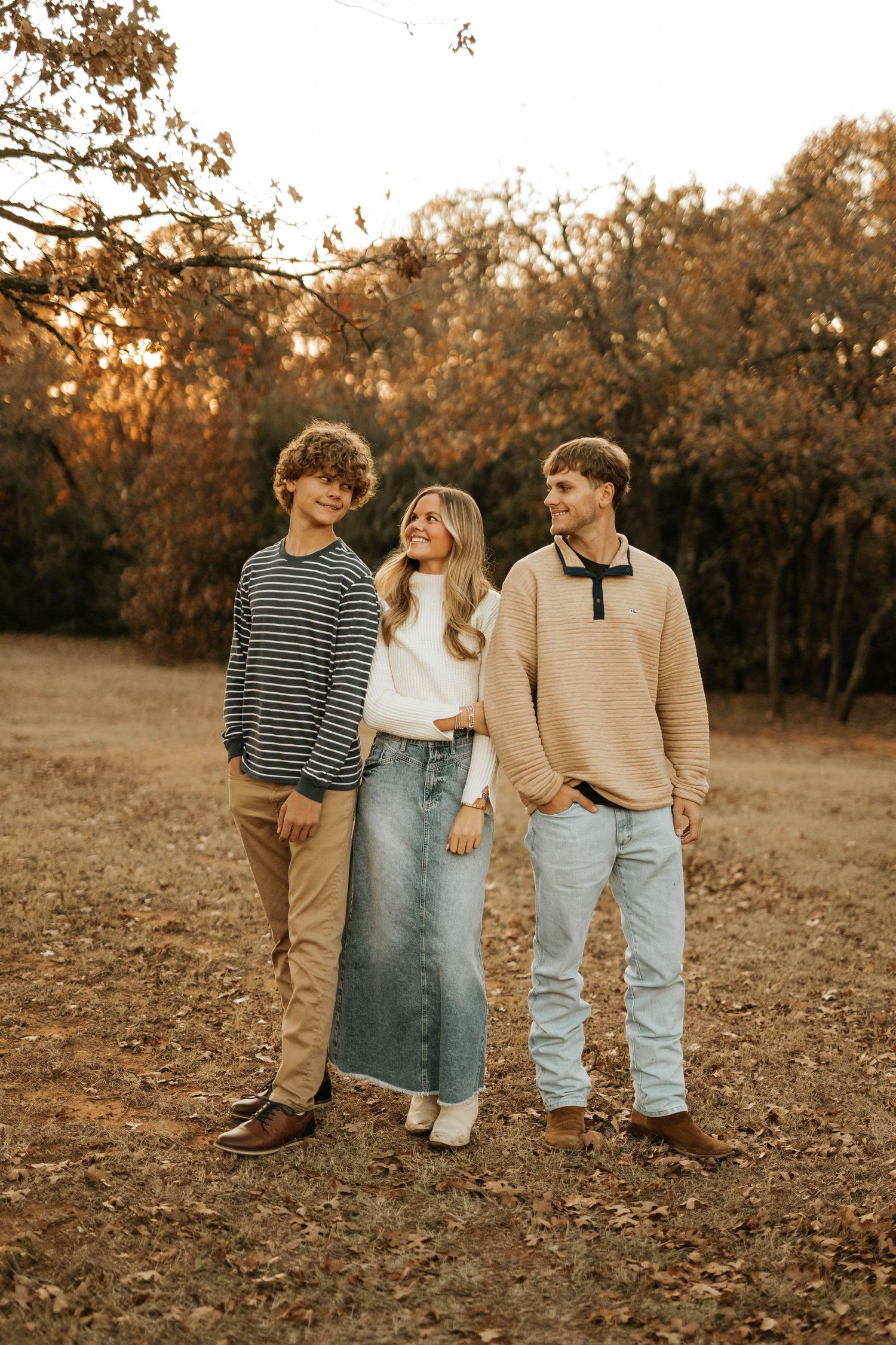 Three friends in cozy fall sweaters pose together in autumn woodland setting.