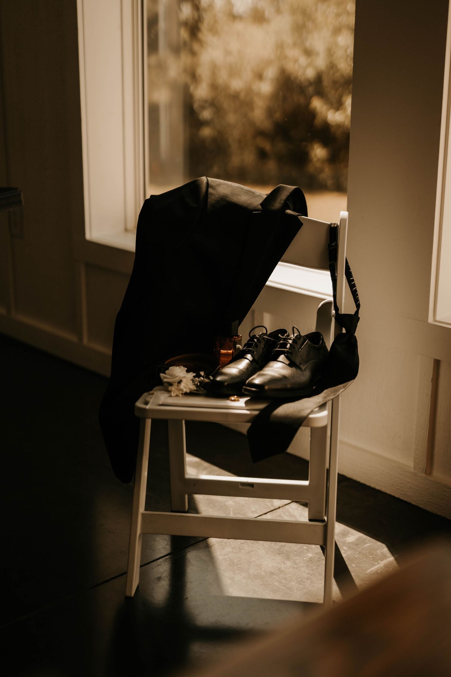 Men's formal black suit and shoes arranged on wooden chair in moody lighting.