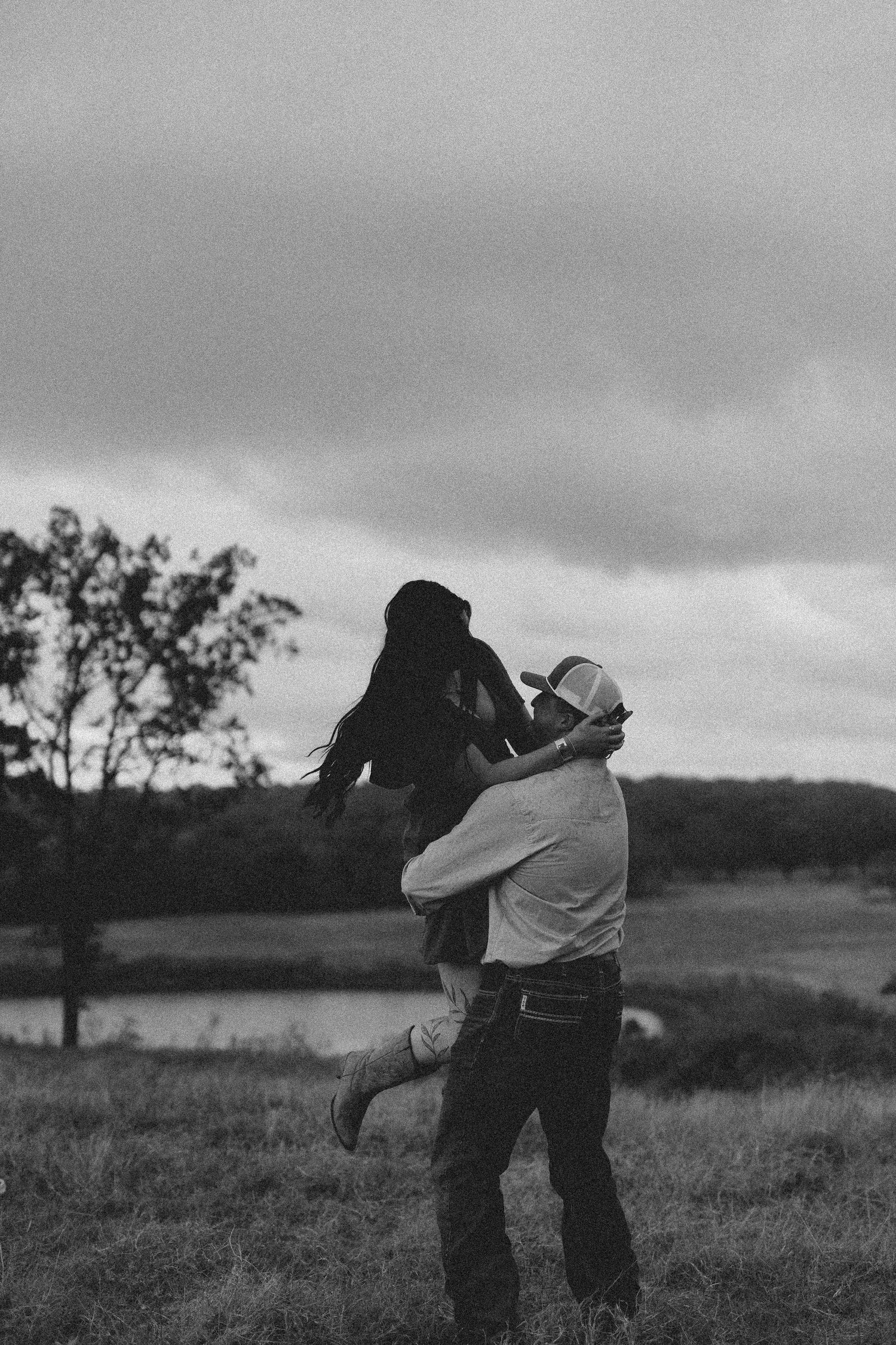 Black and white photo of a man in a cowboy hat lifting a woman in a field with trees and cloudy sky.