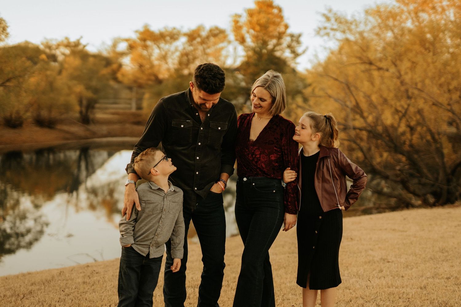 Family of four laughing together outdoors in autumn, standing near a pond with golden fall foliage in background.