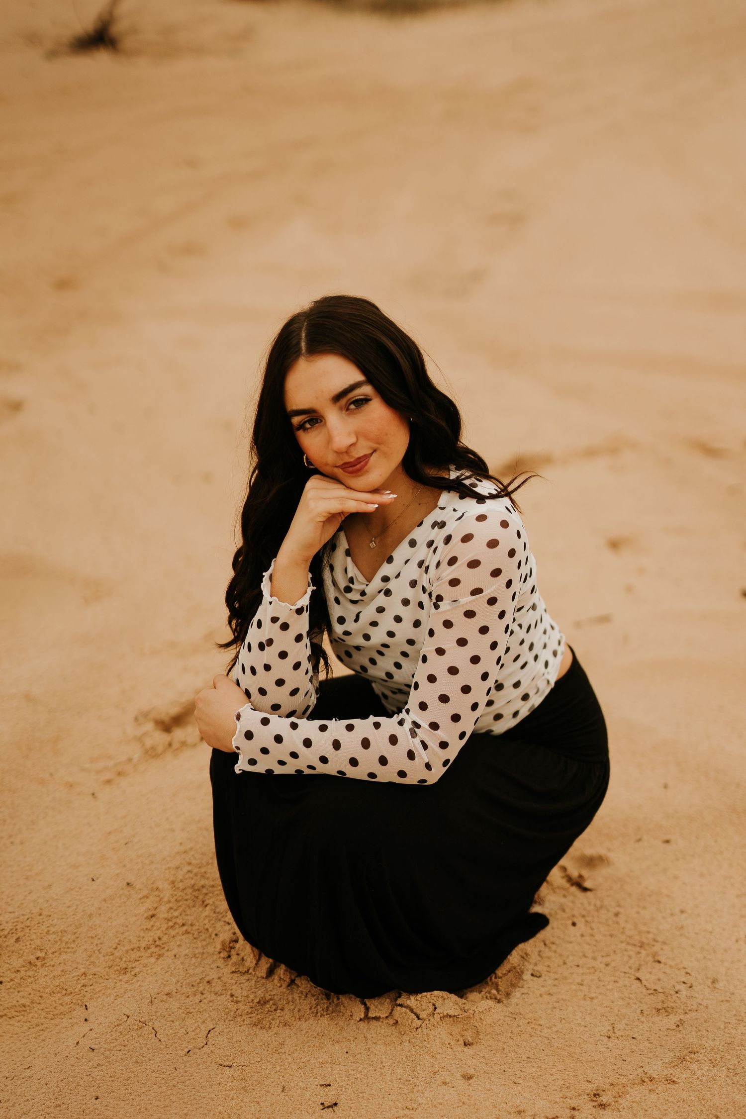 Young woman in polka dot top and black skirt crouching on sandy desert ground, smiling at camera.
