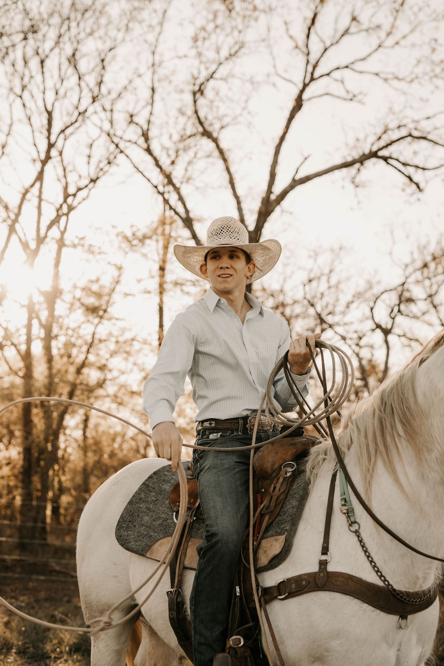 Person in cowboy hat and western attire riding a white horse through wooded area at sunset.