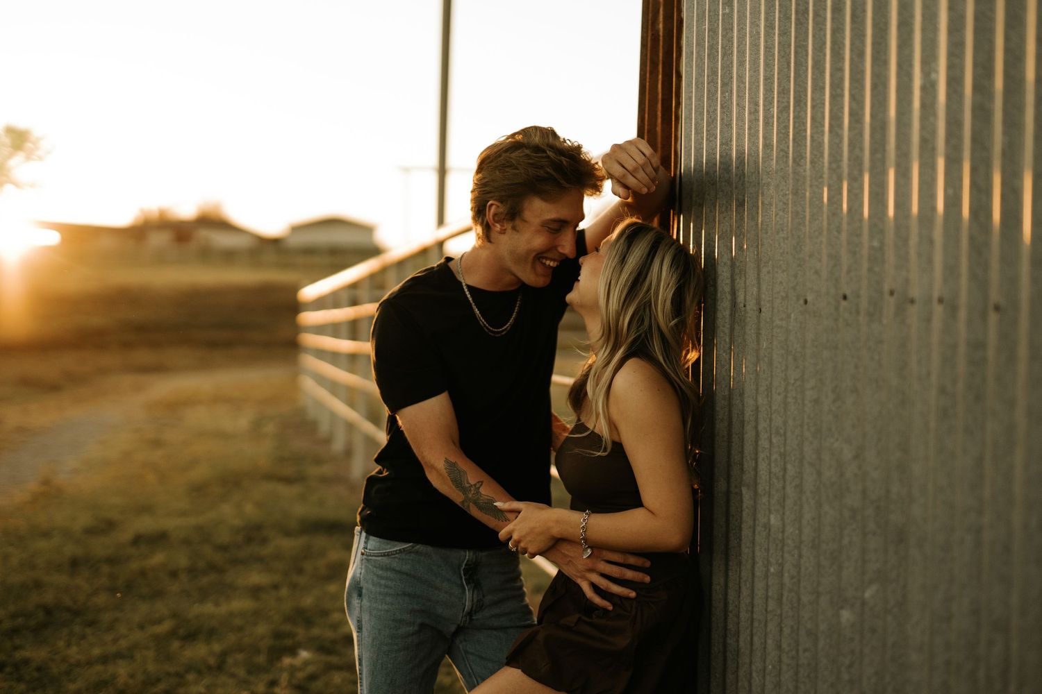 Couple sharing a romantic moment leaning against a rustic metal wall during golden hour sunset on a farm.