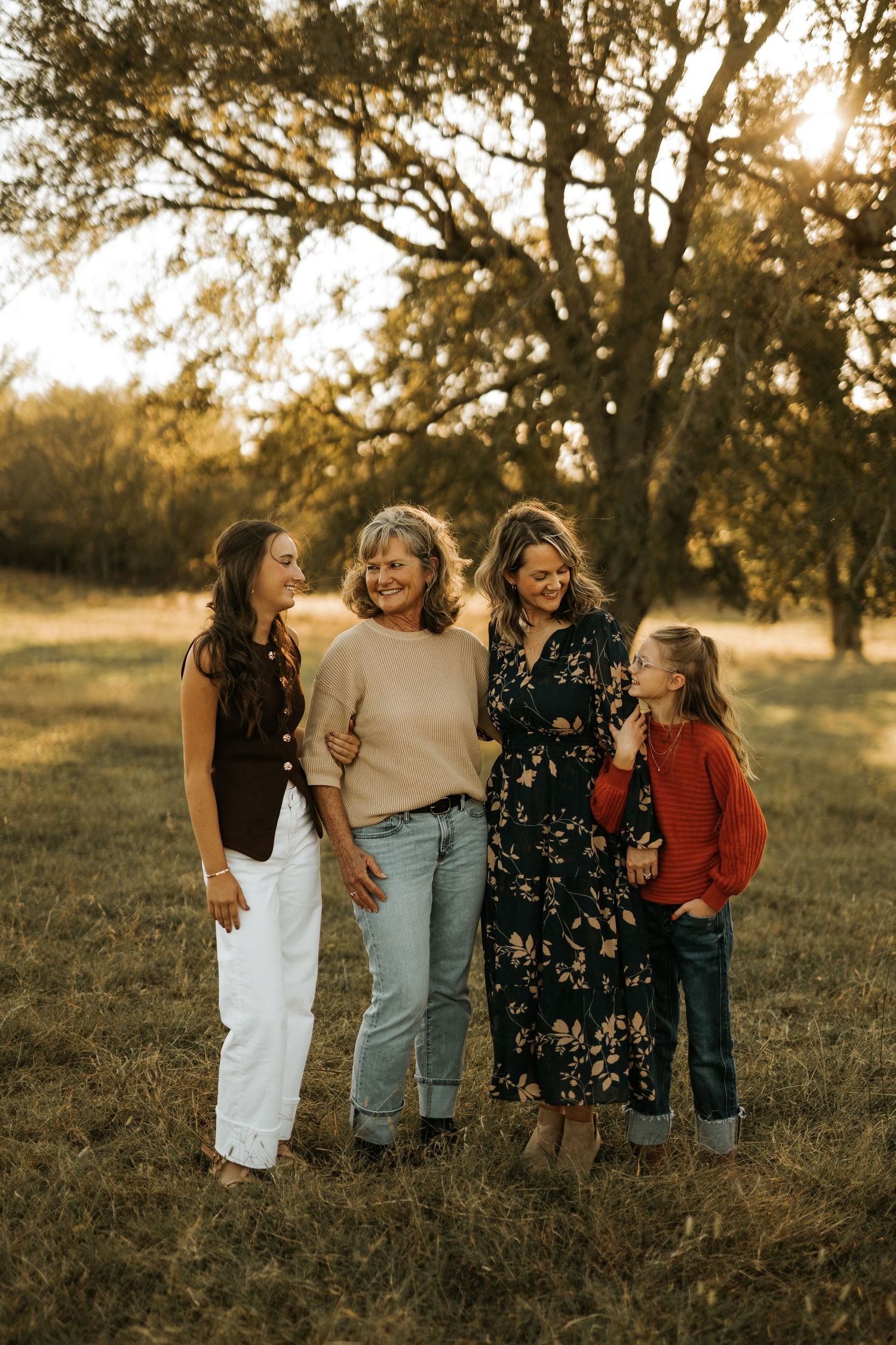 Four women smiling and laughing together in a sunlit field with golden trees in the background during autumn.