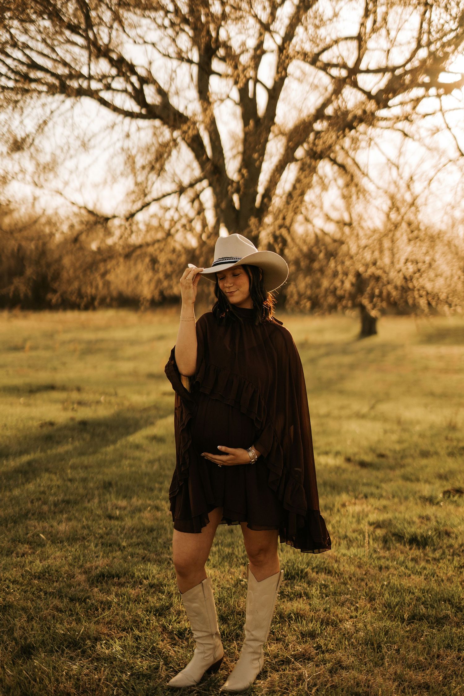 Pregnant woman in black dress, white cowboy hat and boots poses in golden hour sunlight beside a bare tree in a grassy field.