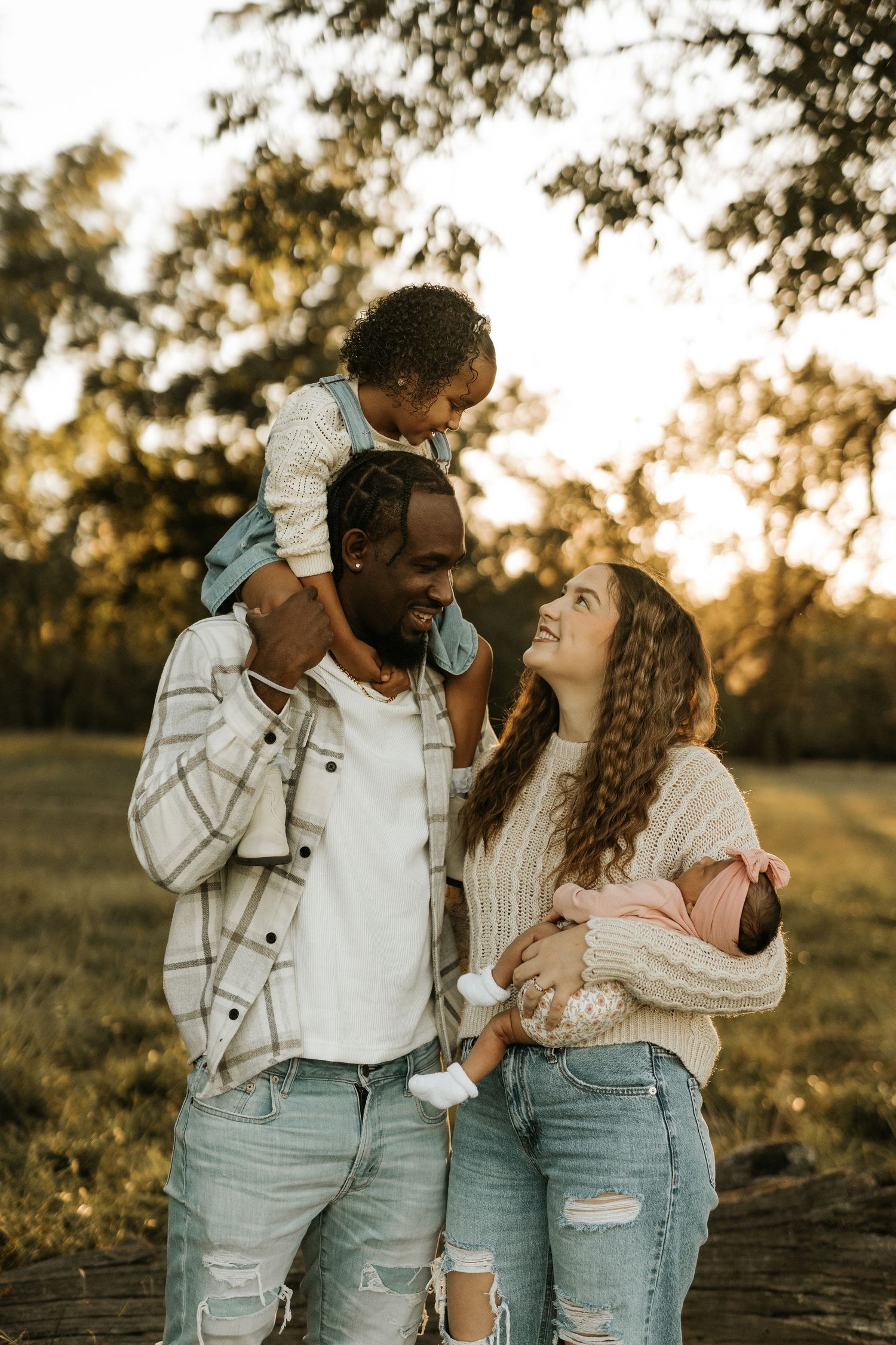A happy interracial family poses outdoors at golden hour, with dad carrying toddler on shoulders and mom holding newborn baby.