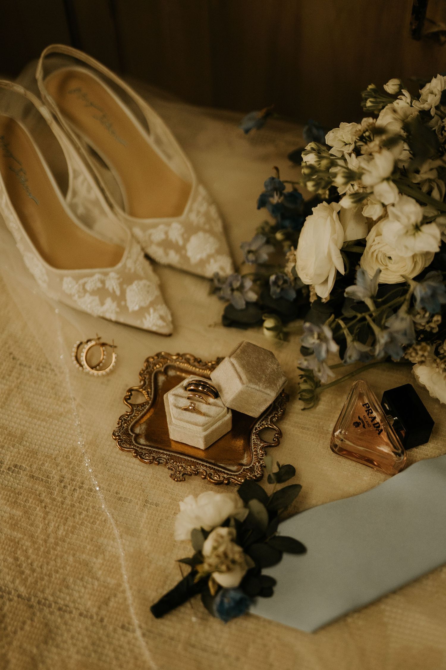 Elegant wedding flat lay featuring lace bridal heels, white and blue bouquet, gold ring box, perfume, and floral boutonniere.