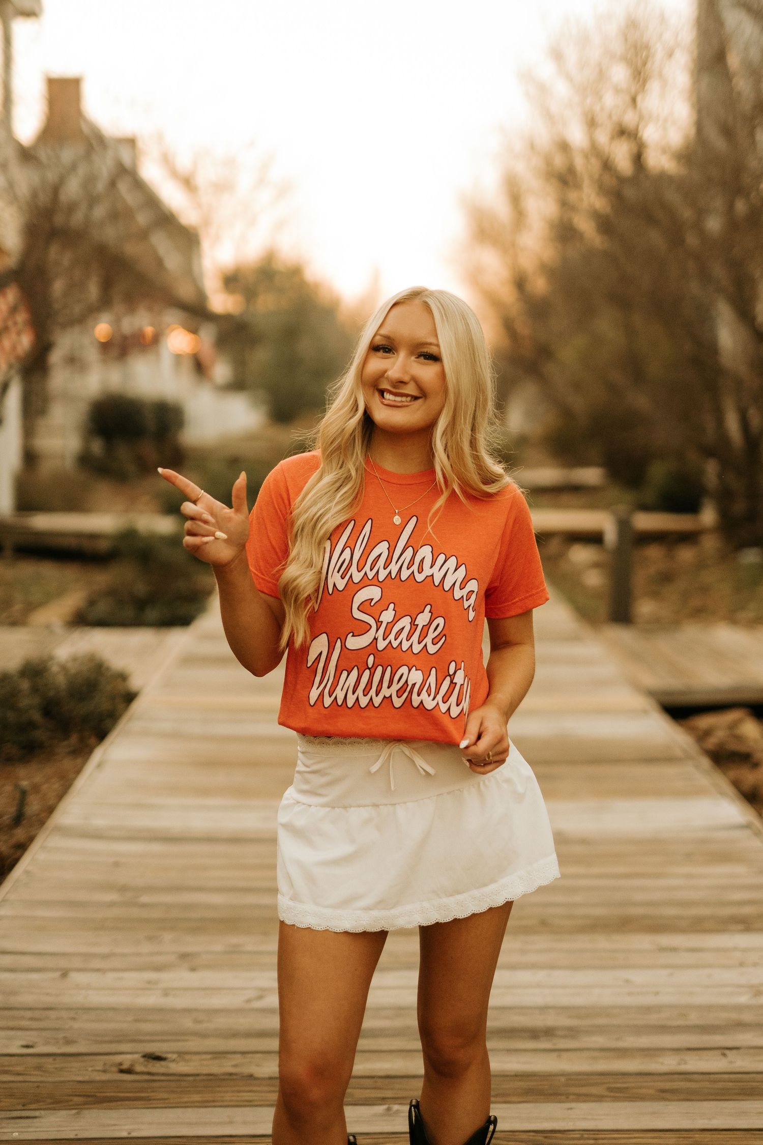 Smiling blonde woman wearing an Oklahoma State University orange t-shirt and white skirt on a wooden boardwalk.