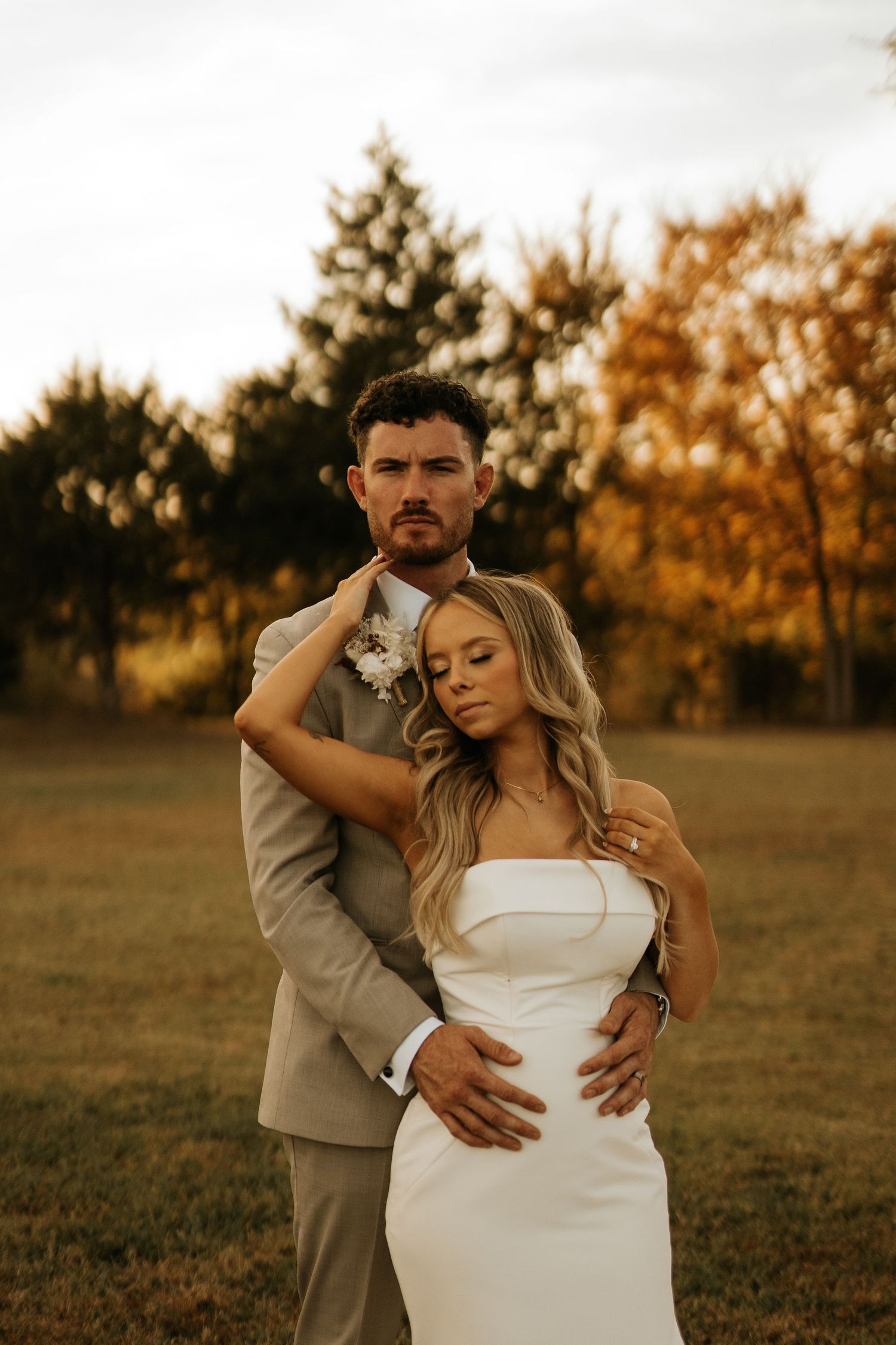 A bride and groom embrace in a golden autumn field, she in a white strapless gown, he in a tan suit with boutonniere.