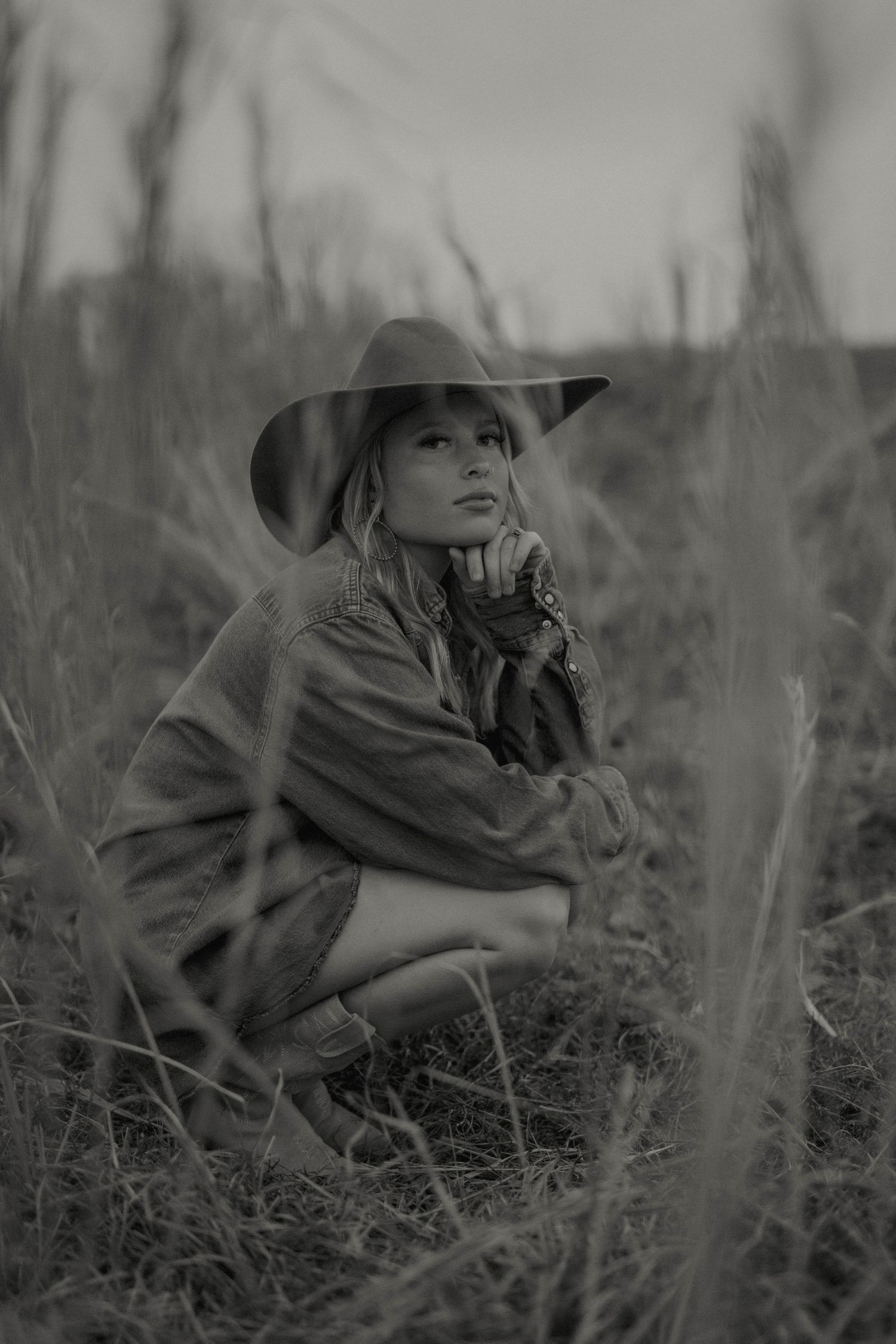 Black and white portrait of a person in a wide brim hat crouching in tall grass with a contemplative expression.