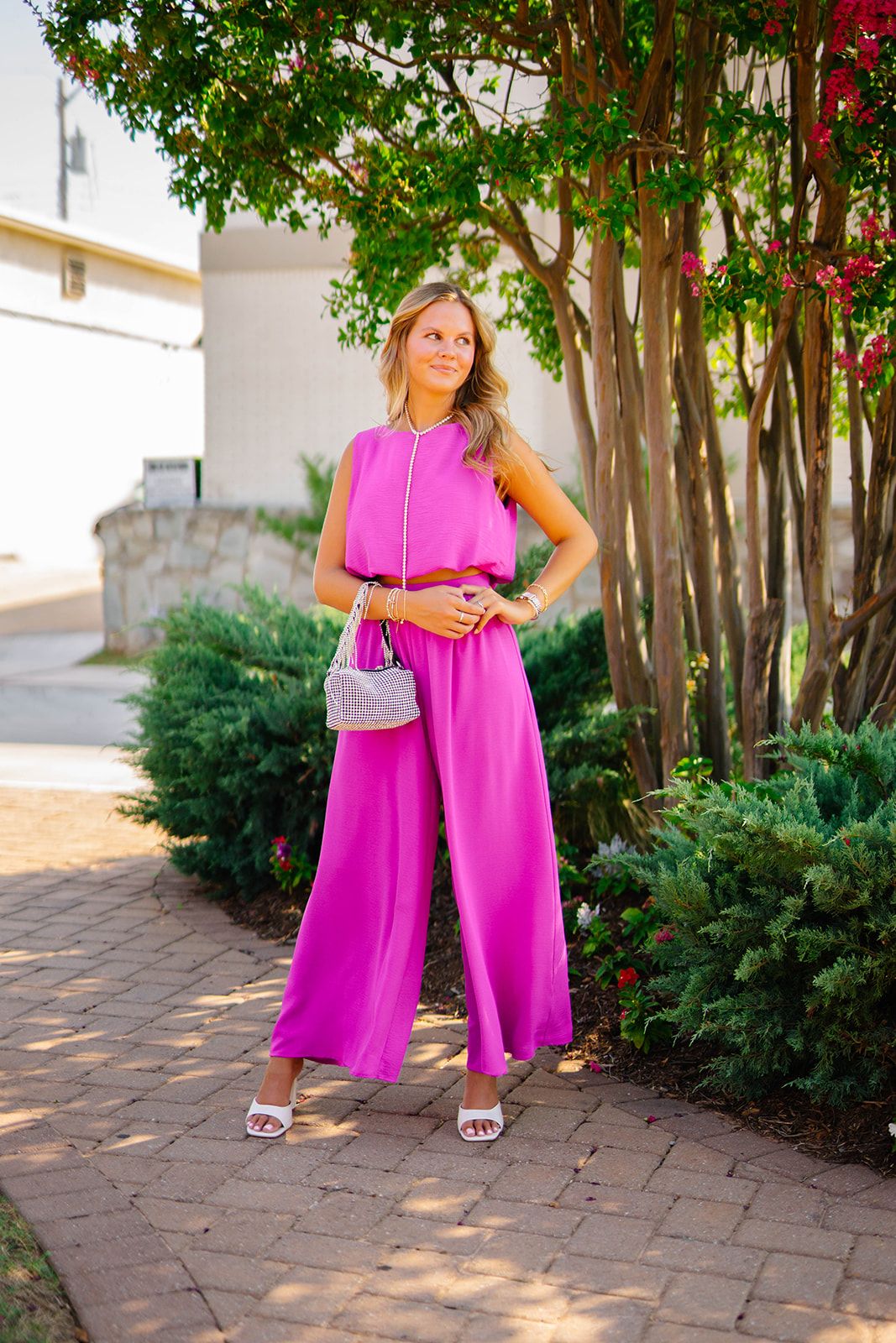 Woman in bright pink jumpsuit poses on brick pathway with trees in background.