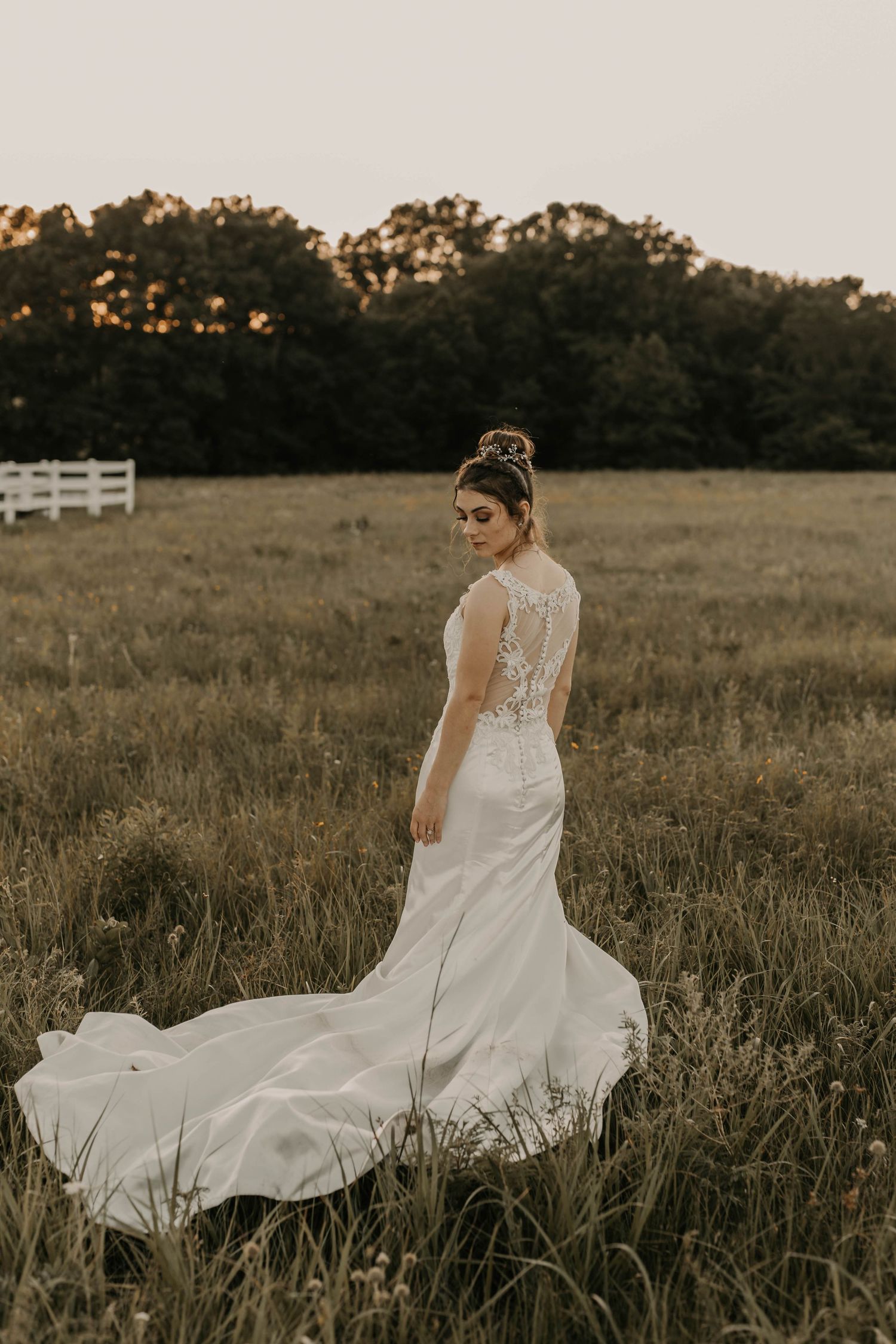 Bride in white lace gown with train poses in golden field at sunset.