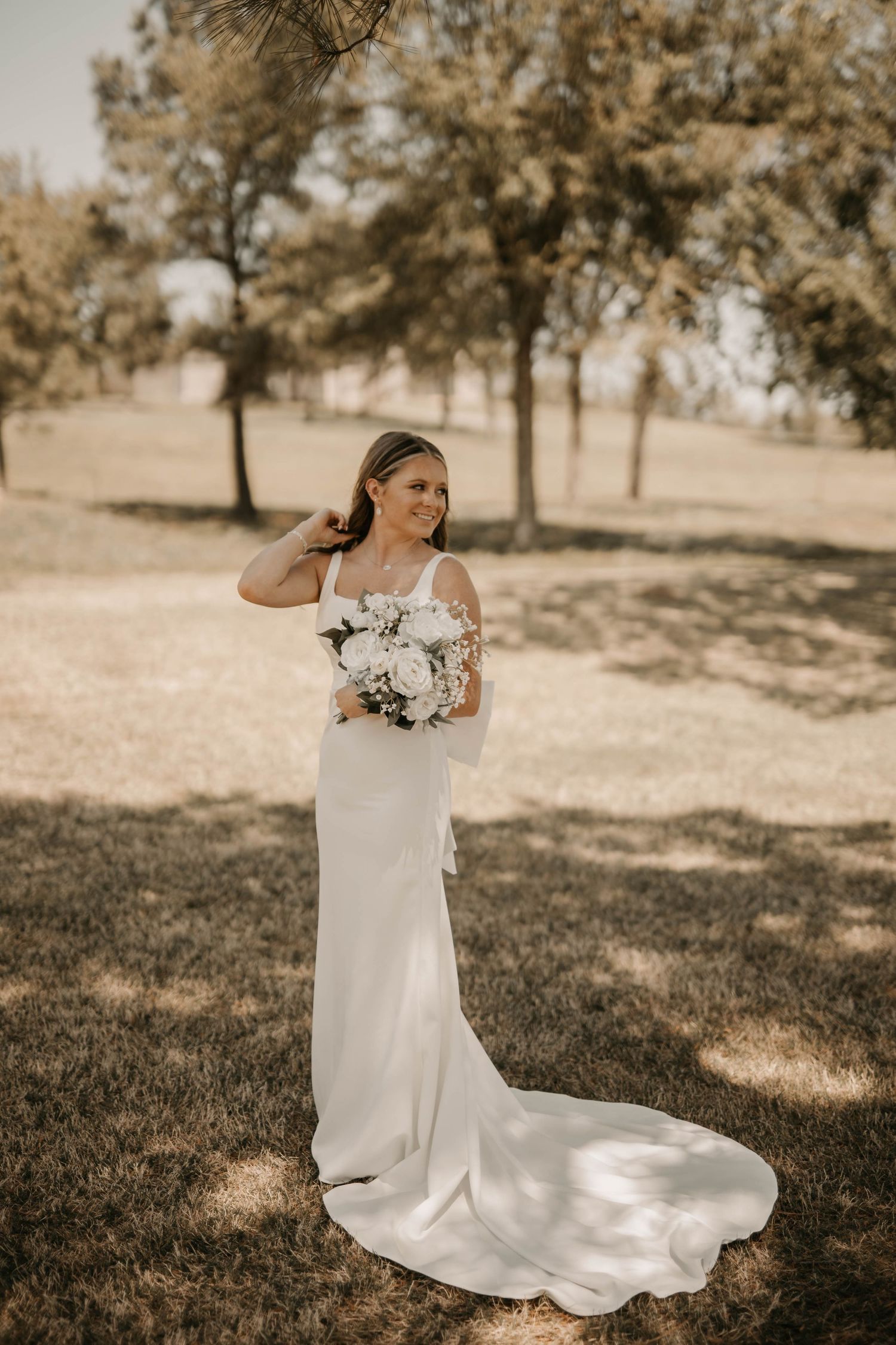 An elegant bride in a fitted white gown holds a bouquet while posing in a sun-dappled outdoor setting.
