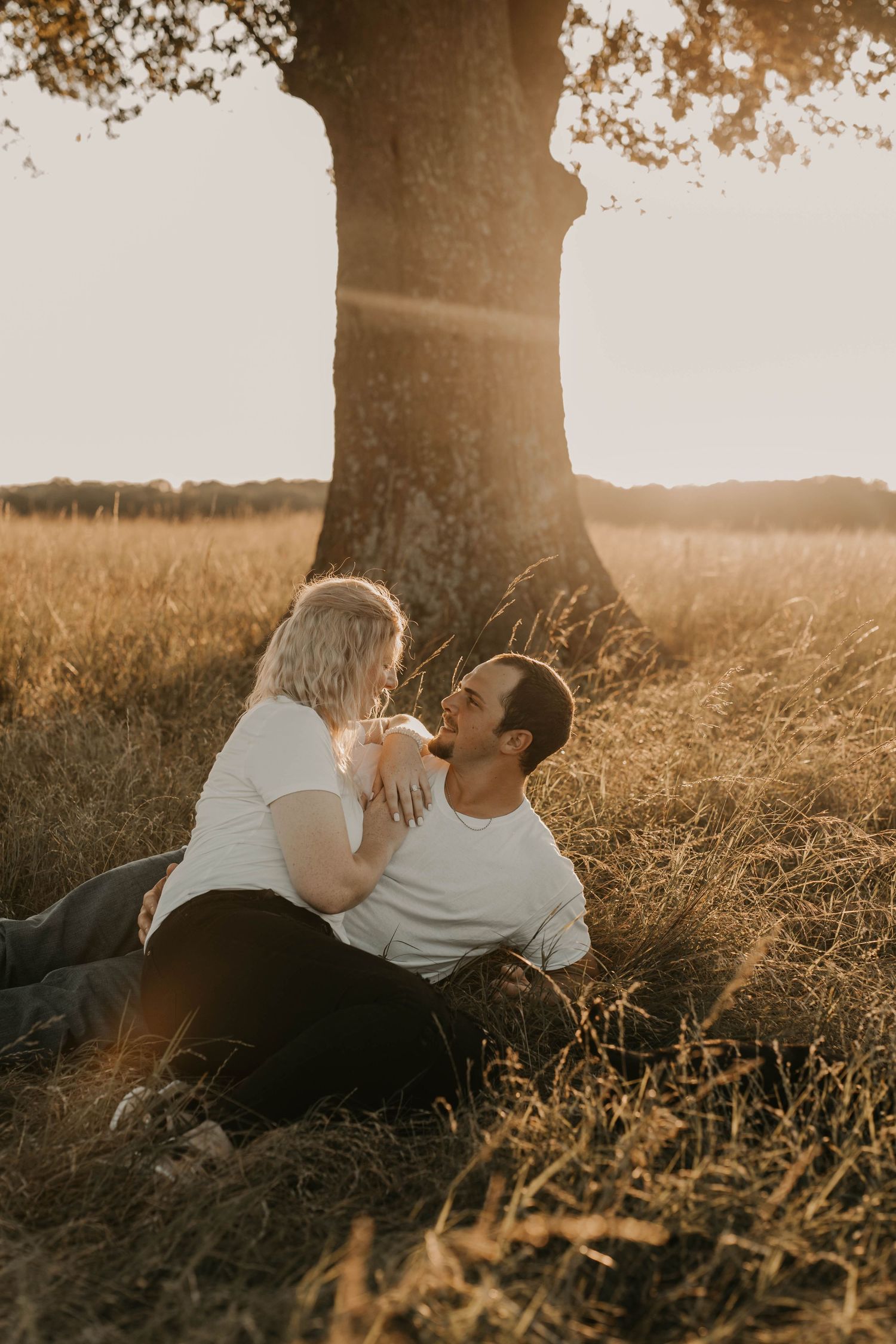Romantic couple sharing an intimate moment under a large tree at golden hour in a grassy field.