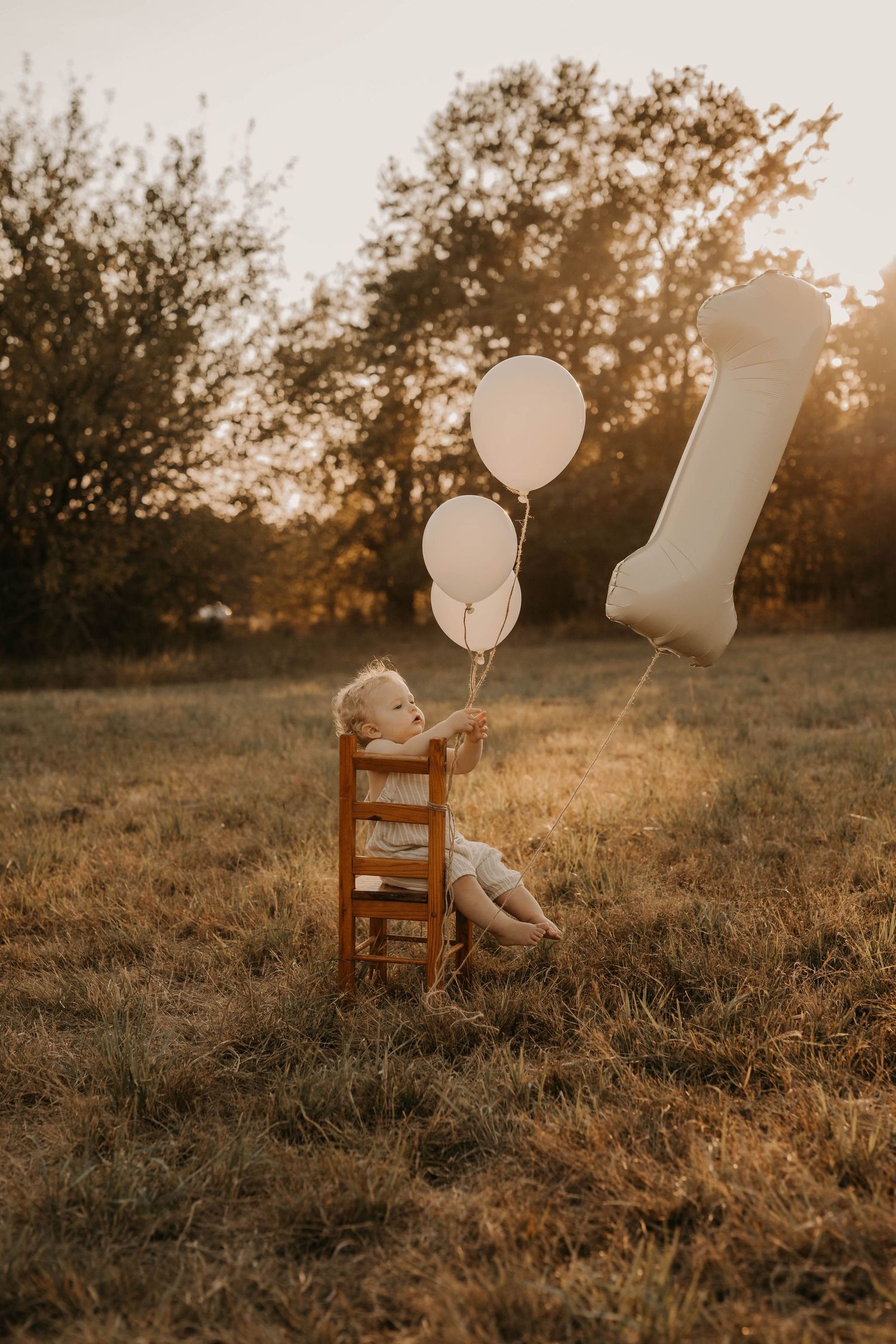 A wooden chair and white balloons sit in a golden field at sunset.