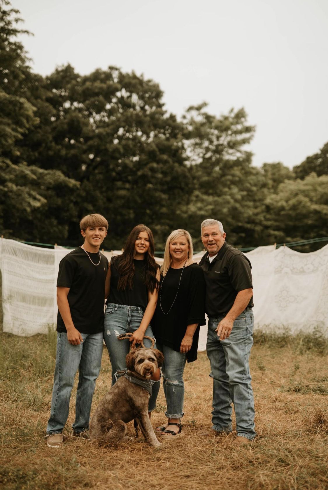 Family of four in matching black tops and jeans posing outdoors with their brown dog near a clothesline.
