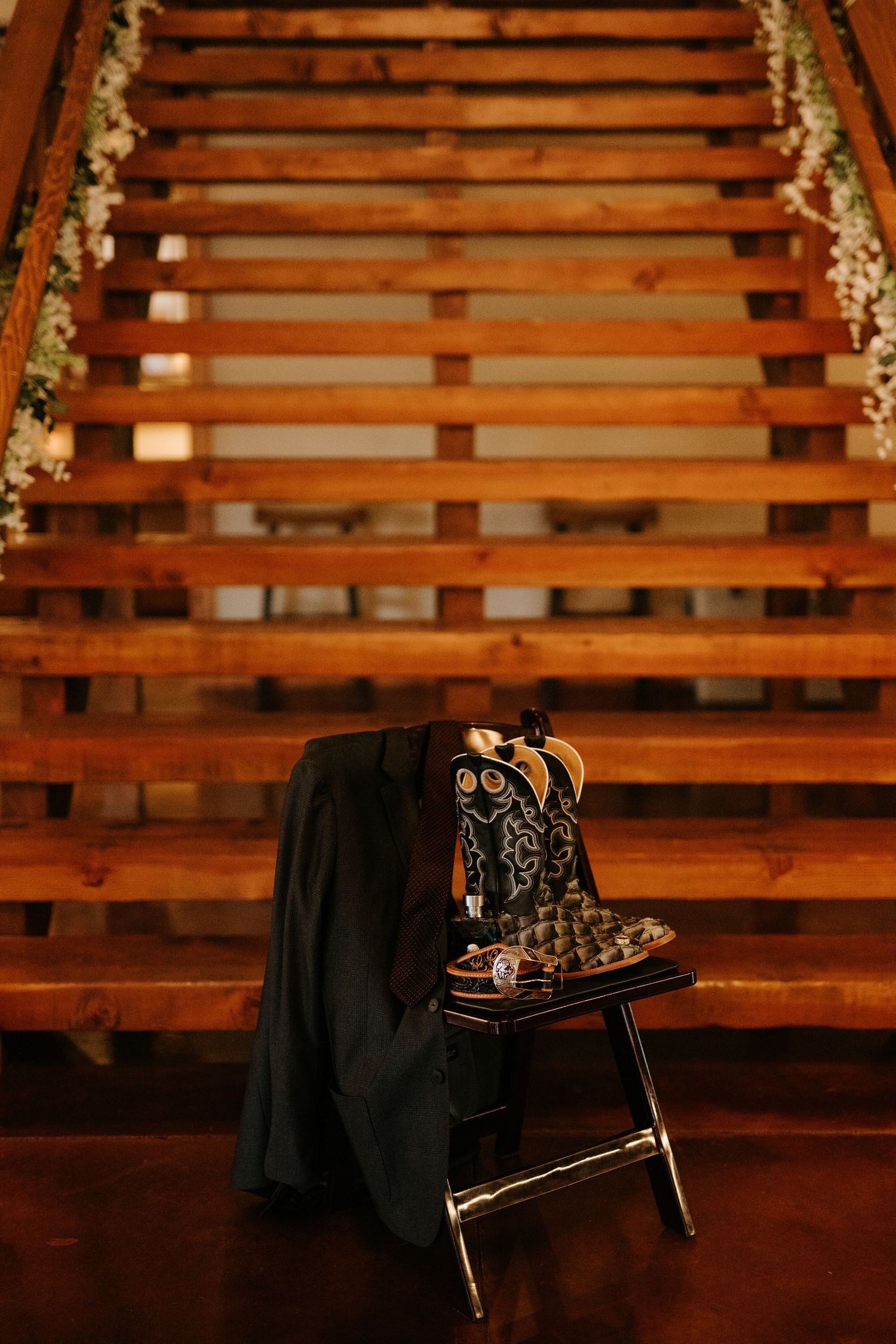 Decorative cowboy boots and black suit jacket displayed on a stool at the base of wooden stairs adorned with floral garland.