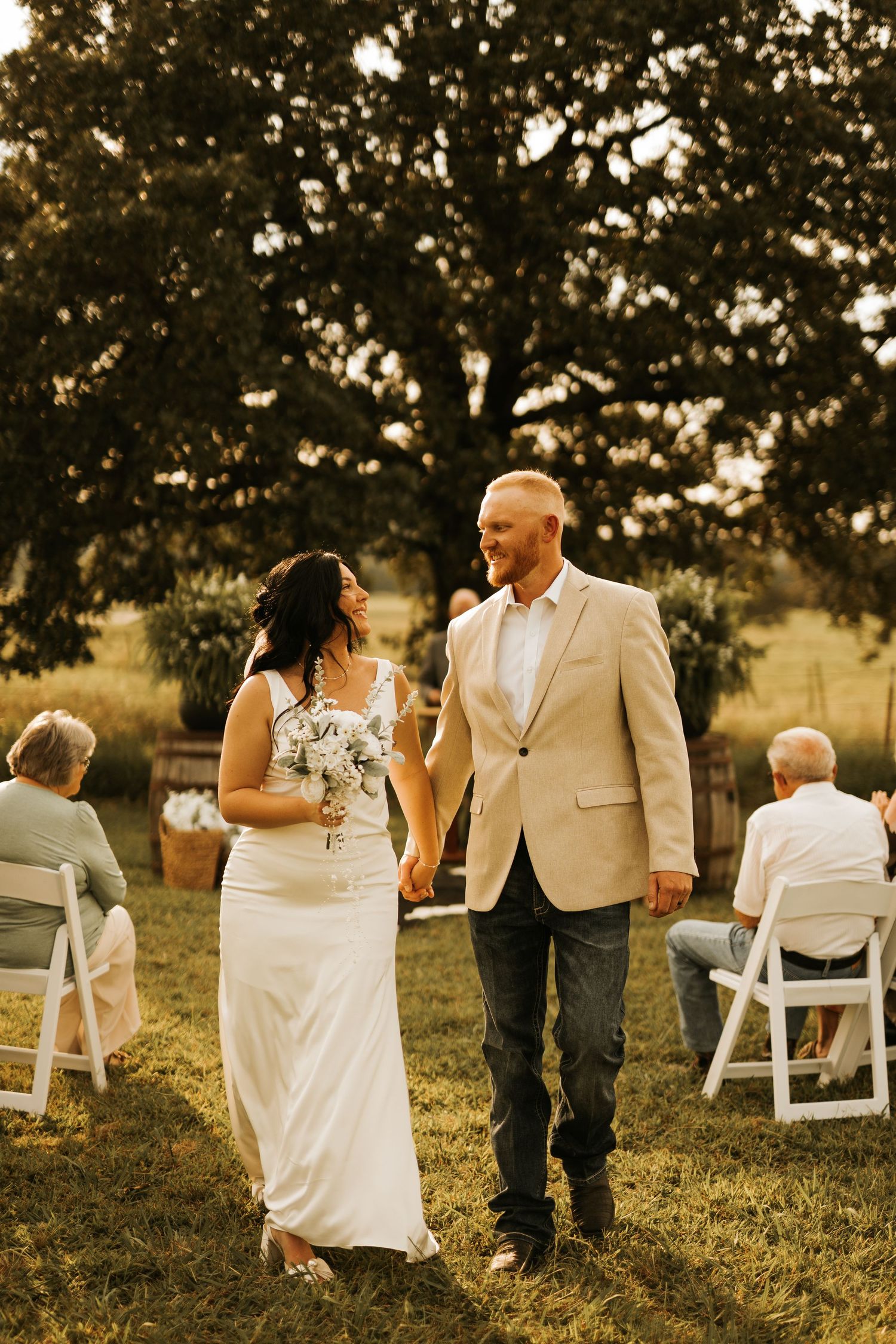 Bride and groom walk hand-in-hand down outdoor aisle after wedding ceremony, guests seated on white chairs under large tree.